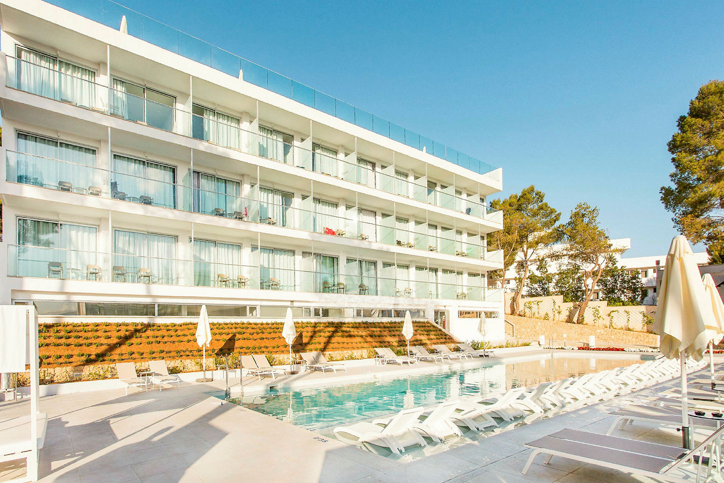 En hotellbygning med flere etasjer og balkonger foran et svømmebasseng omgitt av solsenger og parasoller. (AI-generert tekst)