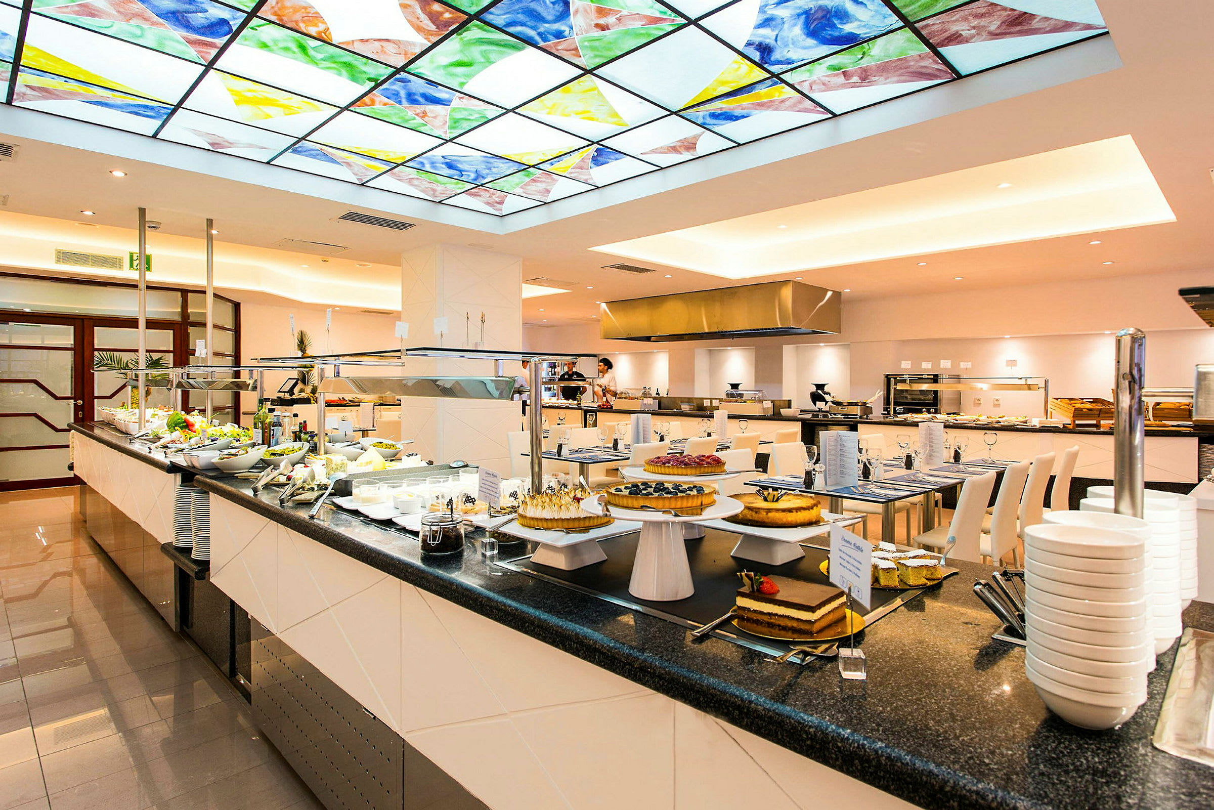 Bildet viser en matbuffé i en restaurant med et fargerikt glasstak over. På bufféen er det ulike retter og desserter lagt frem. (AI-generert tekst)