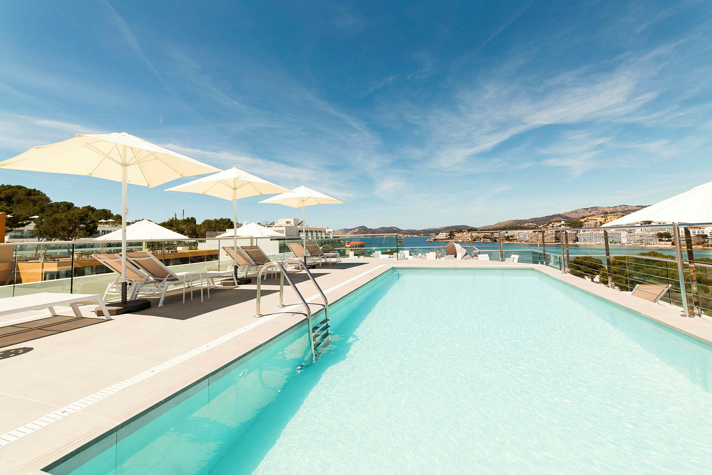 Takterrasse med basseng, solsenger og parasoller, med utsikt over havet og bygninger i bakgrunnen. (AI-generert tekst)