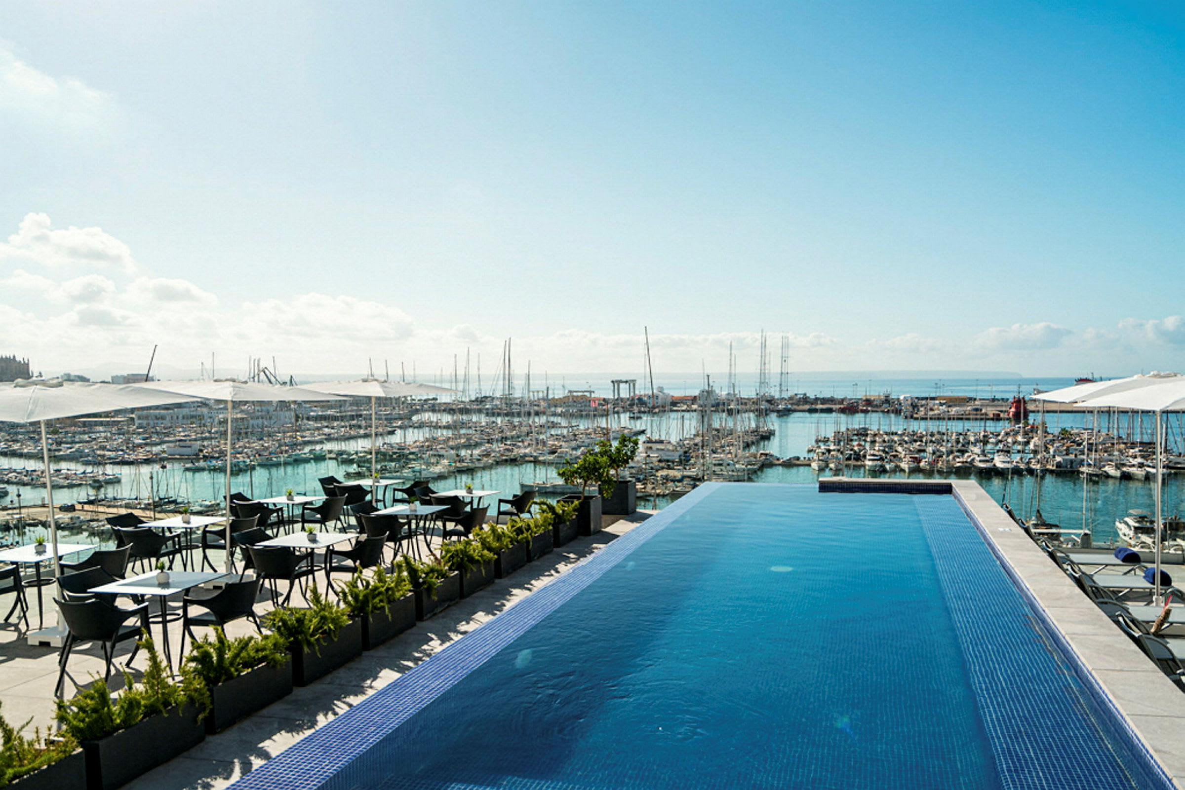 Et infinitybasseng på en terrasse med utsikt over en marina full av båter og havet i bakgrunnen. Det er sitteplasser under parasoller ved siden av bassenget. (AI-generert tekst)
