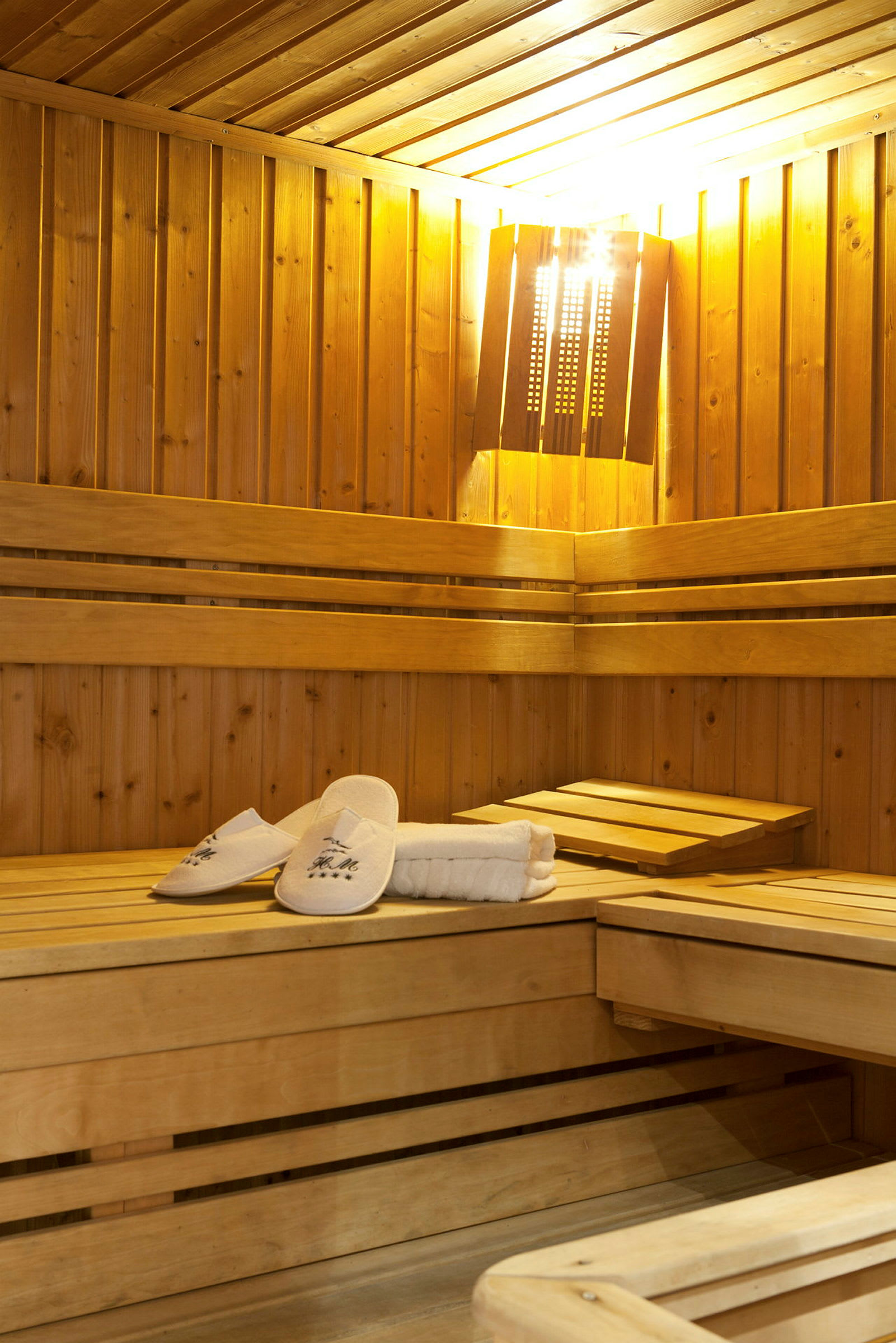 Et tremøblert badstuerom med benker og en mykt belyst lampe. På en benk ligger hvite sandaler og håndklær. (AI-generert tekst)