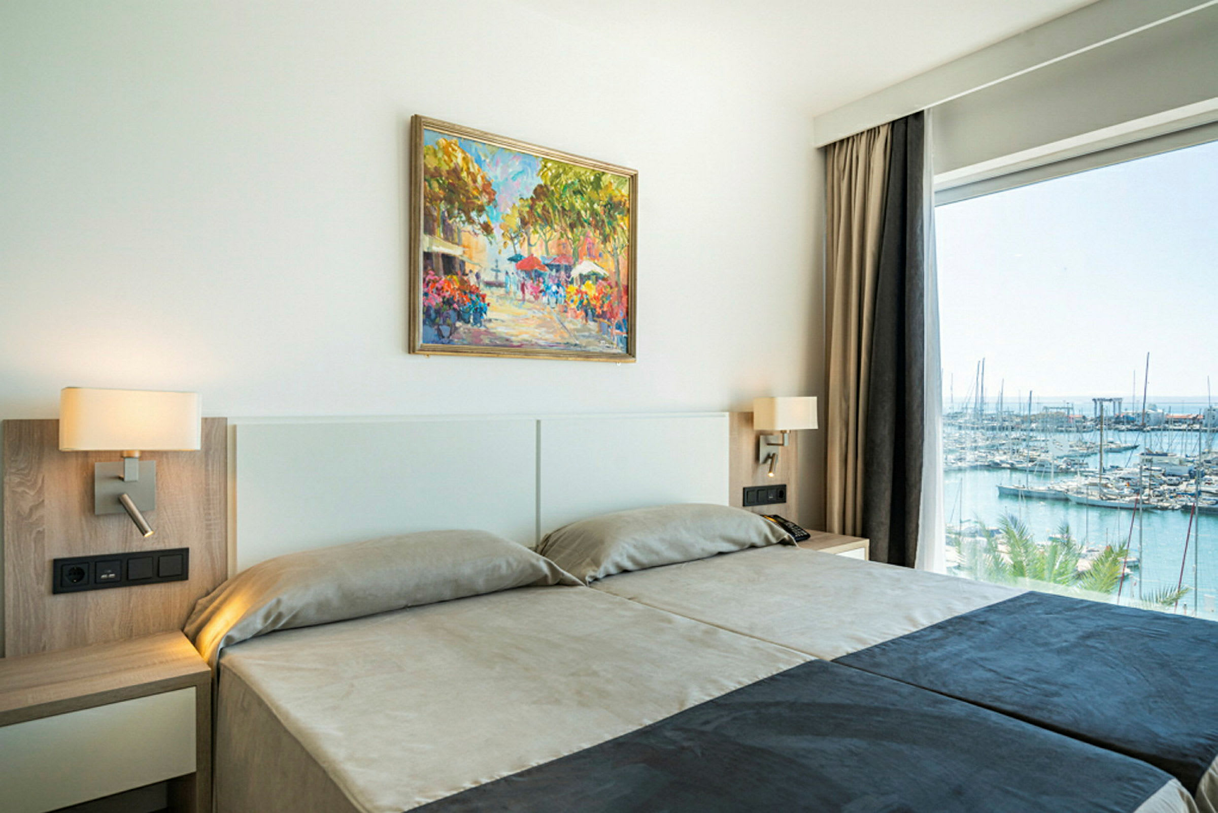 Et hotellrom med to senger, vegglamper, et fargerikt maleri på veggen og en stor vindusåpning med utsikt over en marina. (AI-generert tekst)
