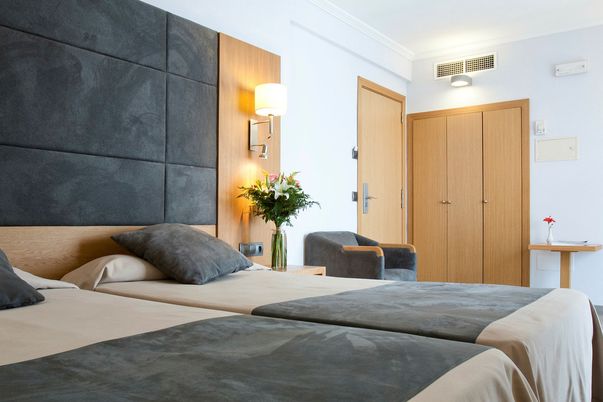 Bilde av et moderne hotellrom med to senger, grå sengetepper, en stol, et tregarderobeskap og en blomsterbukett på et bord. (AI-generert tekst)