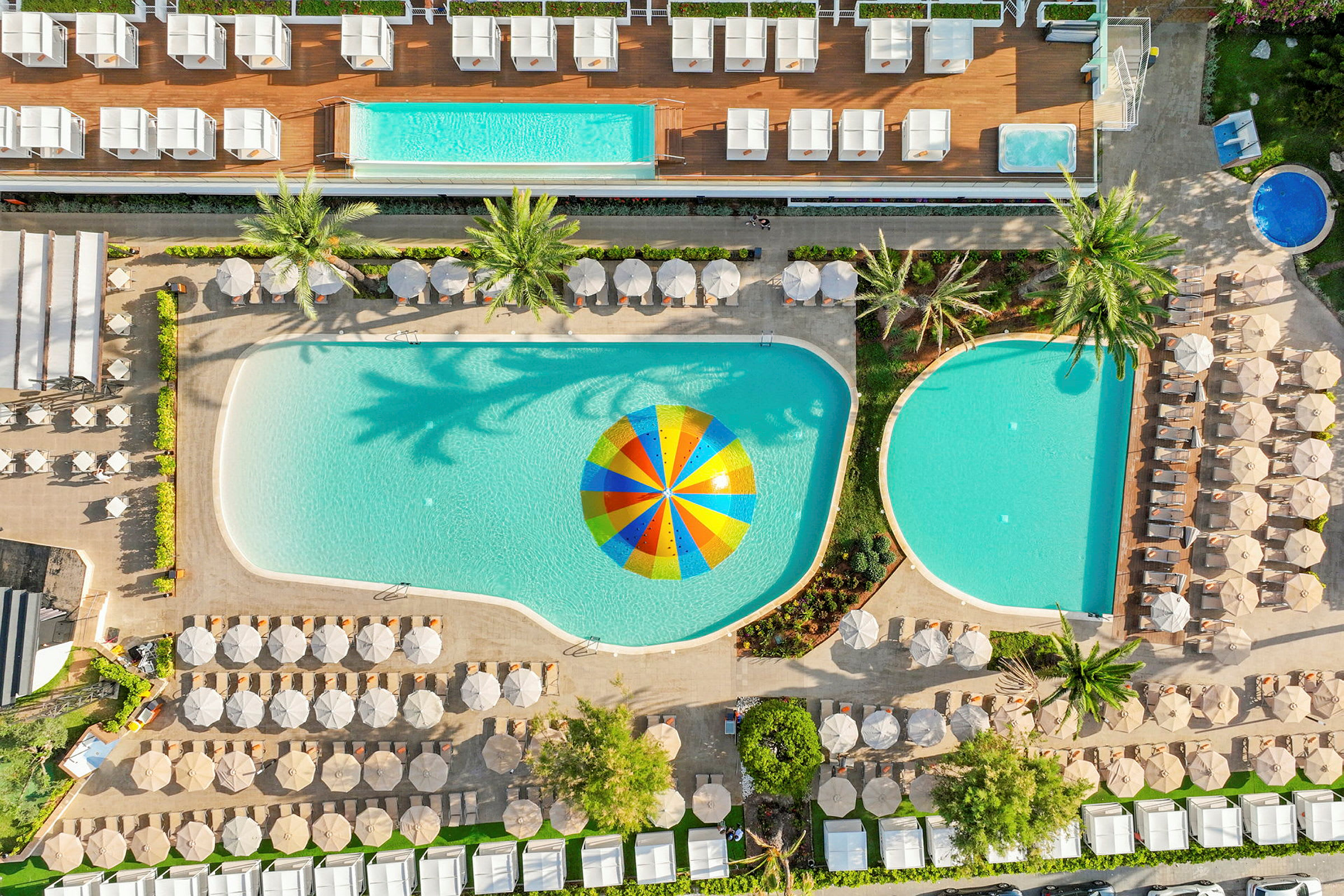 Luftfoto av et hotellbasseng med parasoller og solsenger rundt, samt en fargerik parasoll i vannet. (AI-generert tekst)