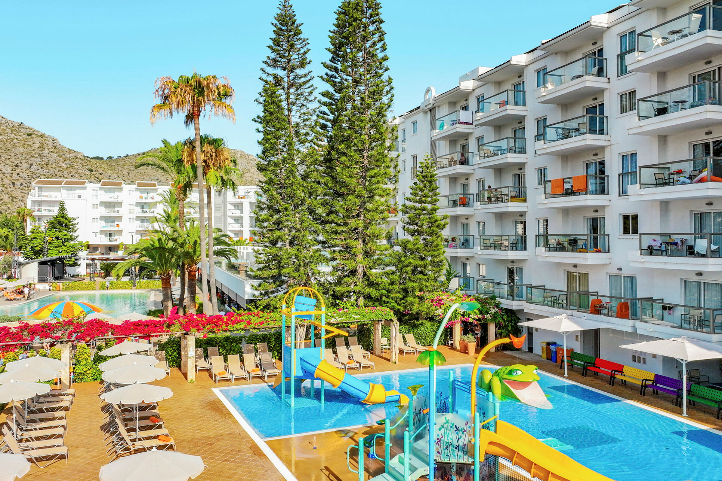 Utsikt over hotellbassengområde med vannsklie, solsenger og omkringliggende bygninger. (AI-generert tekst)