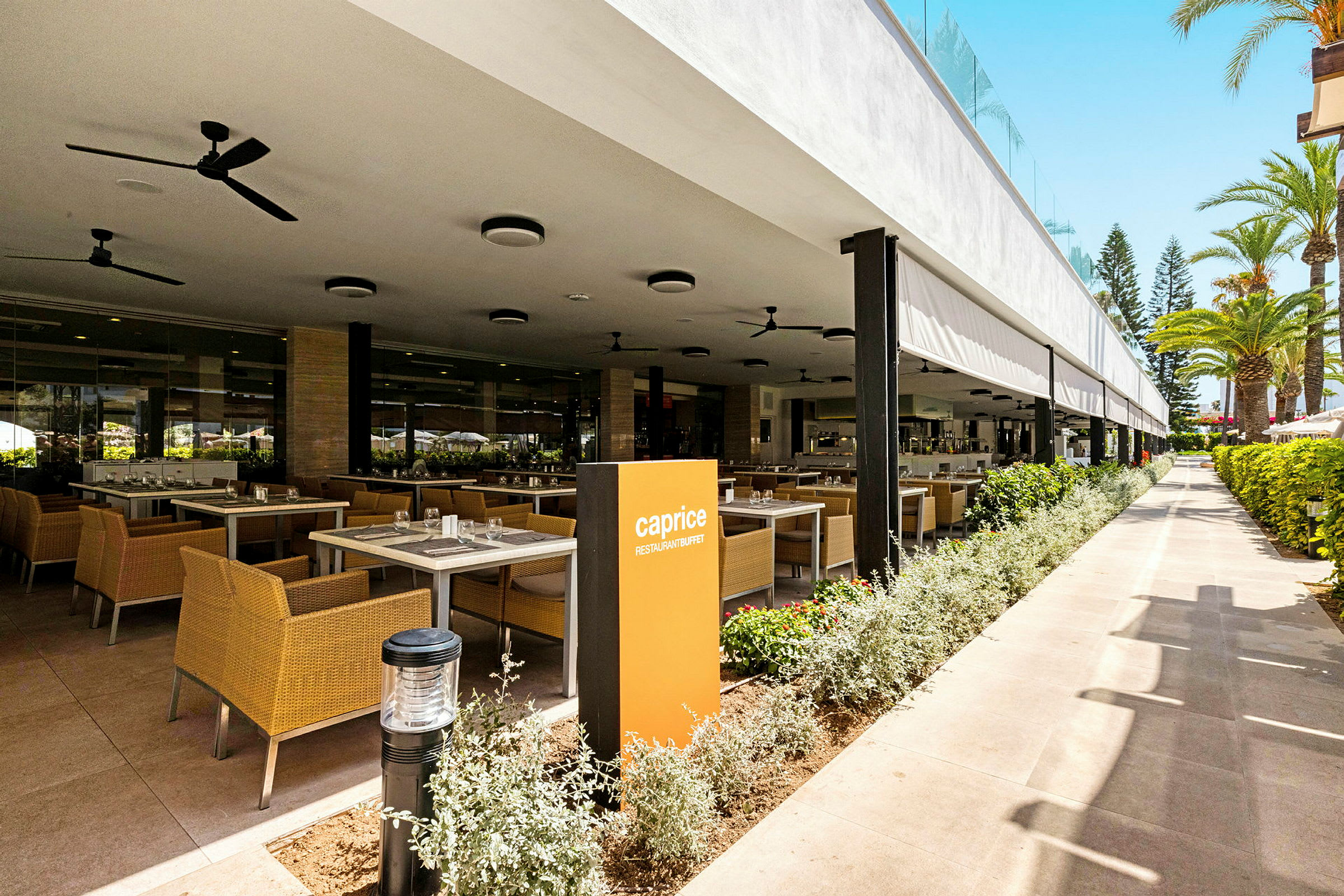 Restaurant med uteservering og bord på rekke ved siden av planter. (AI-generert tekst)