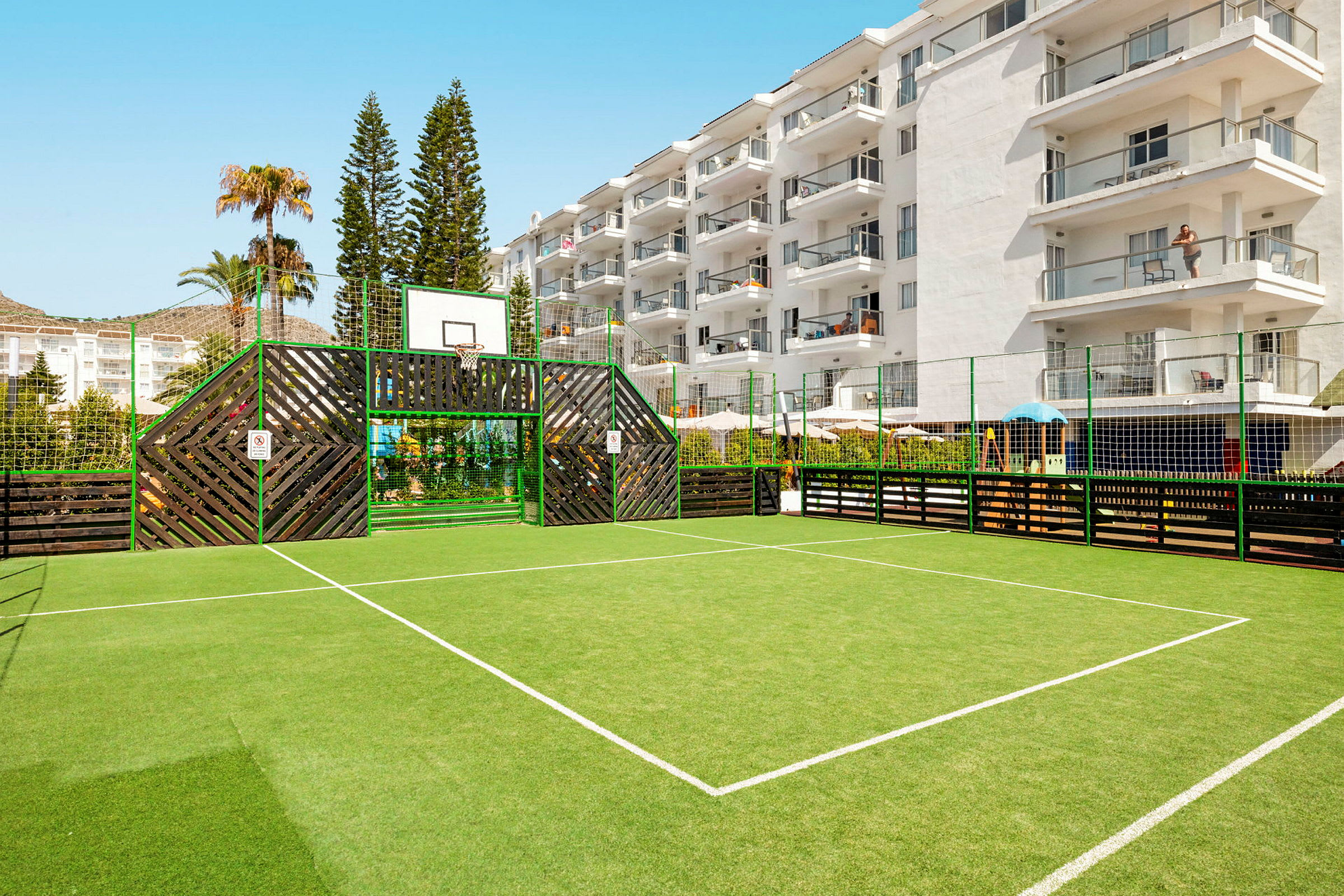 Fotballbane omgitt av gjerde og en hotellbygning i bakgrunnen. (AI-generert tekst)