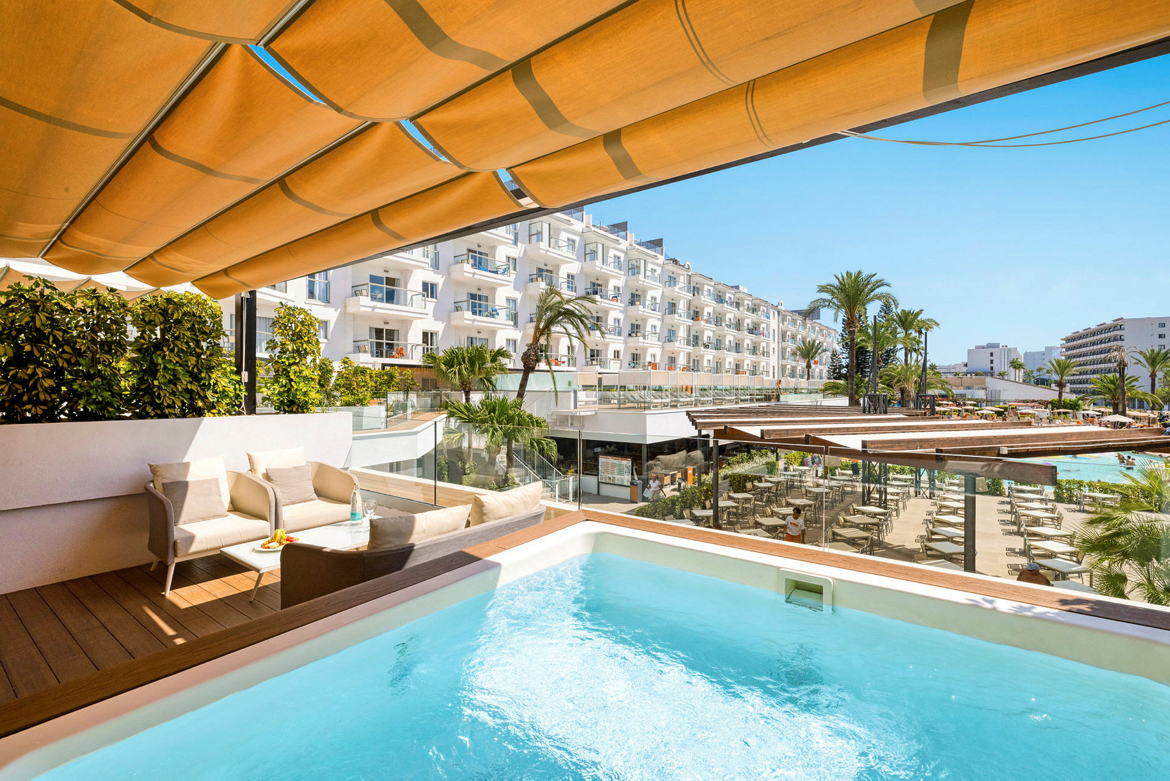 Terrasse med basseng, sittegruppe og utsikt over hotellområde med palmer og bygninger. (AI-generert tekst)