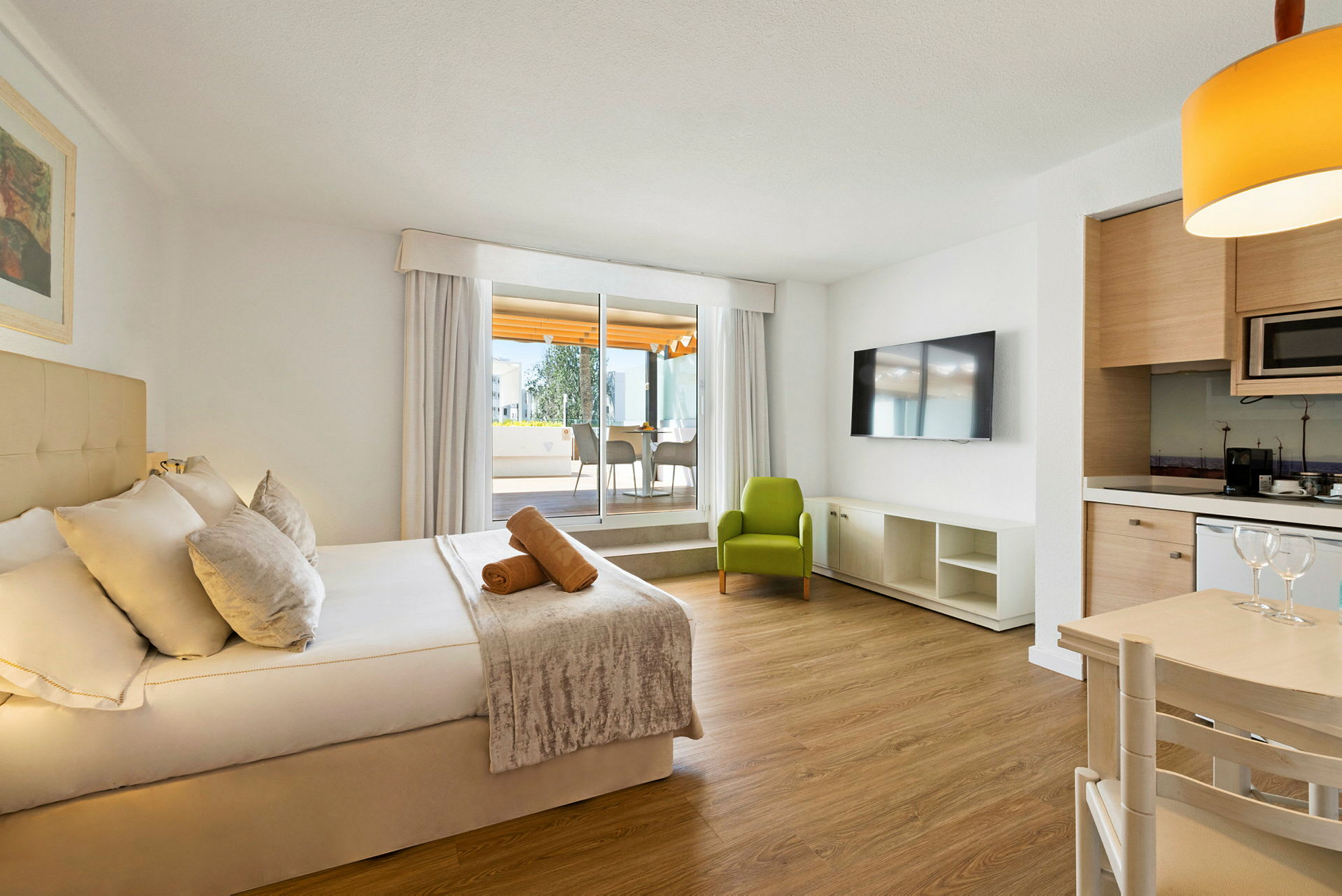 Et moderne hotellrom med en dobbeltseng, en grønn lenestol, en TV og et bord med kjøkkenområde. (AI-generert tekst)