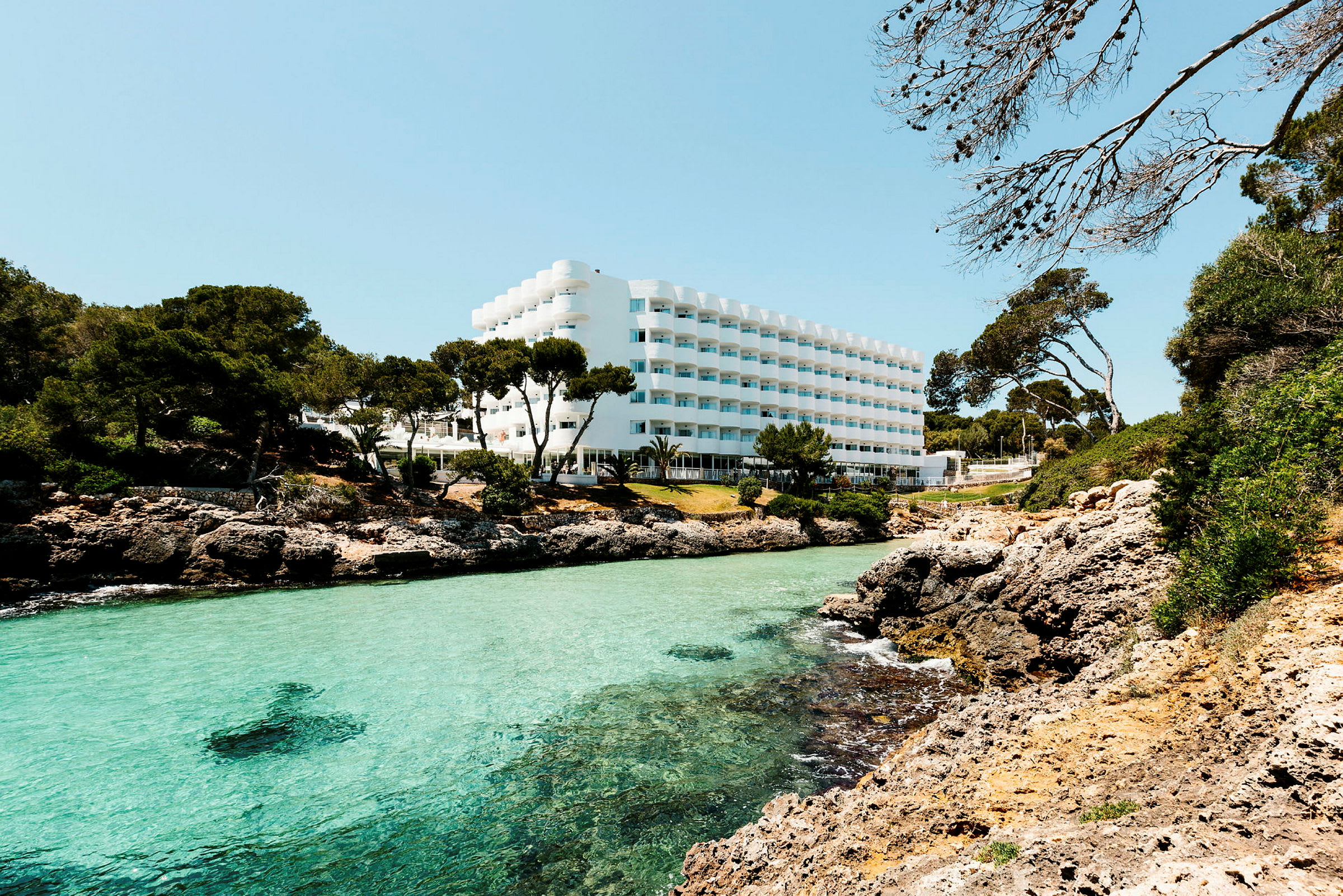 Hotellbygning med hvit fasade, beliggende ved en klipperik kyst med klart turkis vann og omkringliggende grøntområder. (AI-generert tekst)