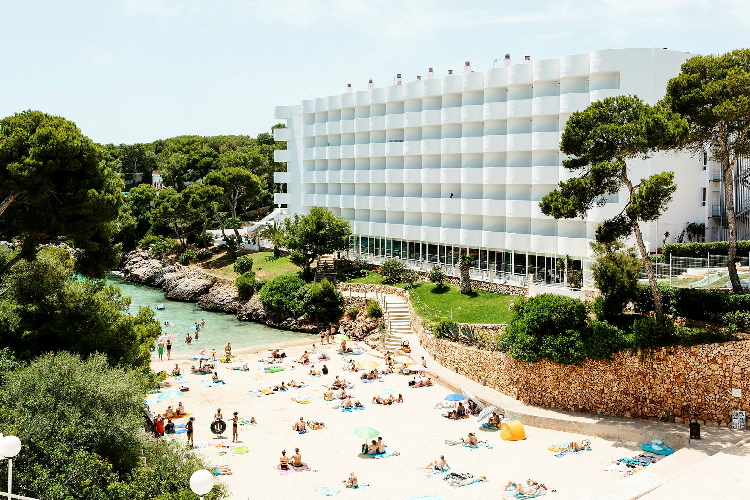 Strand med svømmende mennesker, omgitt av grøntområder og et hvitt hotell. (AI-generert tekst)