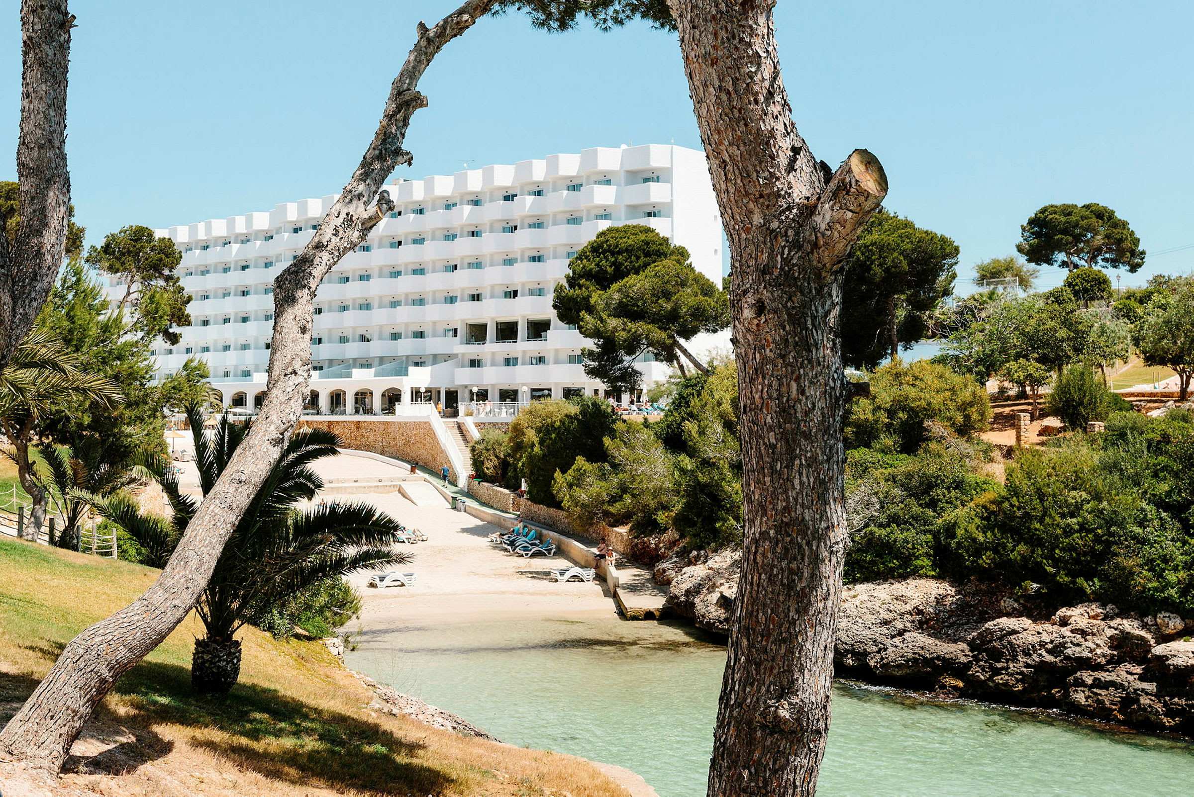 Hotellbygning plassert ved en liten strand, omgitt av trær og grøntområder. (AI-generert tekst)