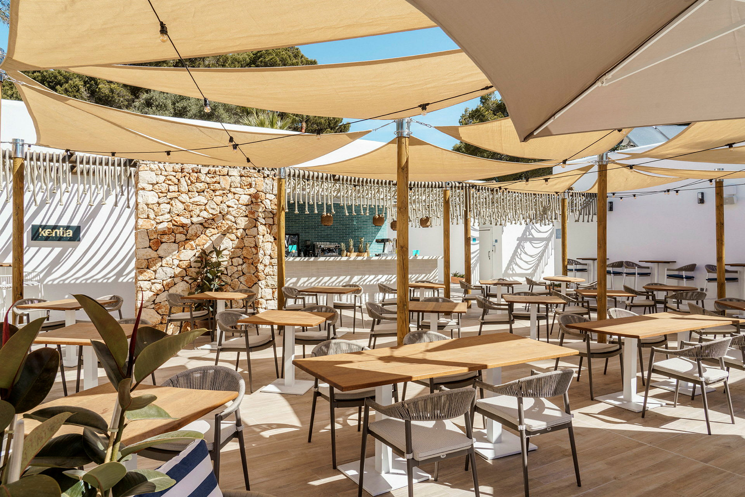 En utendørs restaurant med bord og stoler under skyggefulle seil. (AI-generert tekst)