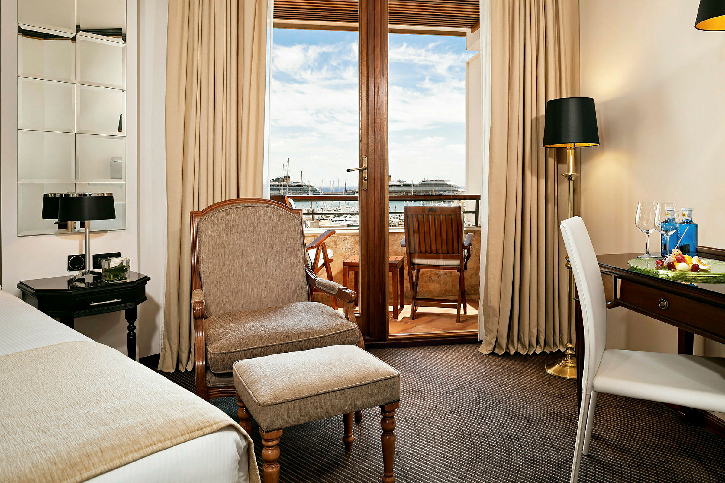 Et hotellrom med en lenestol, et skrivebord og utsikt over en balkong med sitteplass samt en havn i bakgrunnen. (AI-generert tekst)