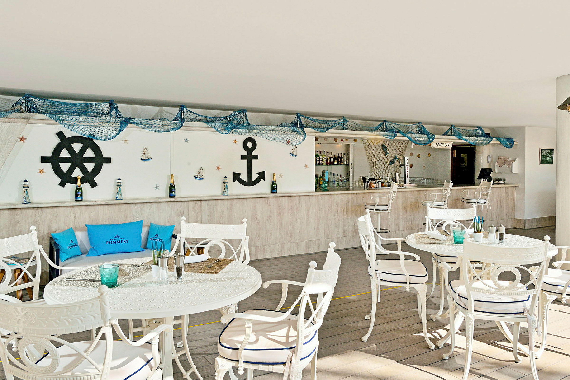 Bilde av en strandbar med maritim innredning, inkludert hvite bord og stoler, blå puter og dekorasjoner som anker og fiskegarn. (AI-generert tekst)