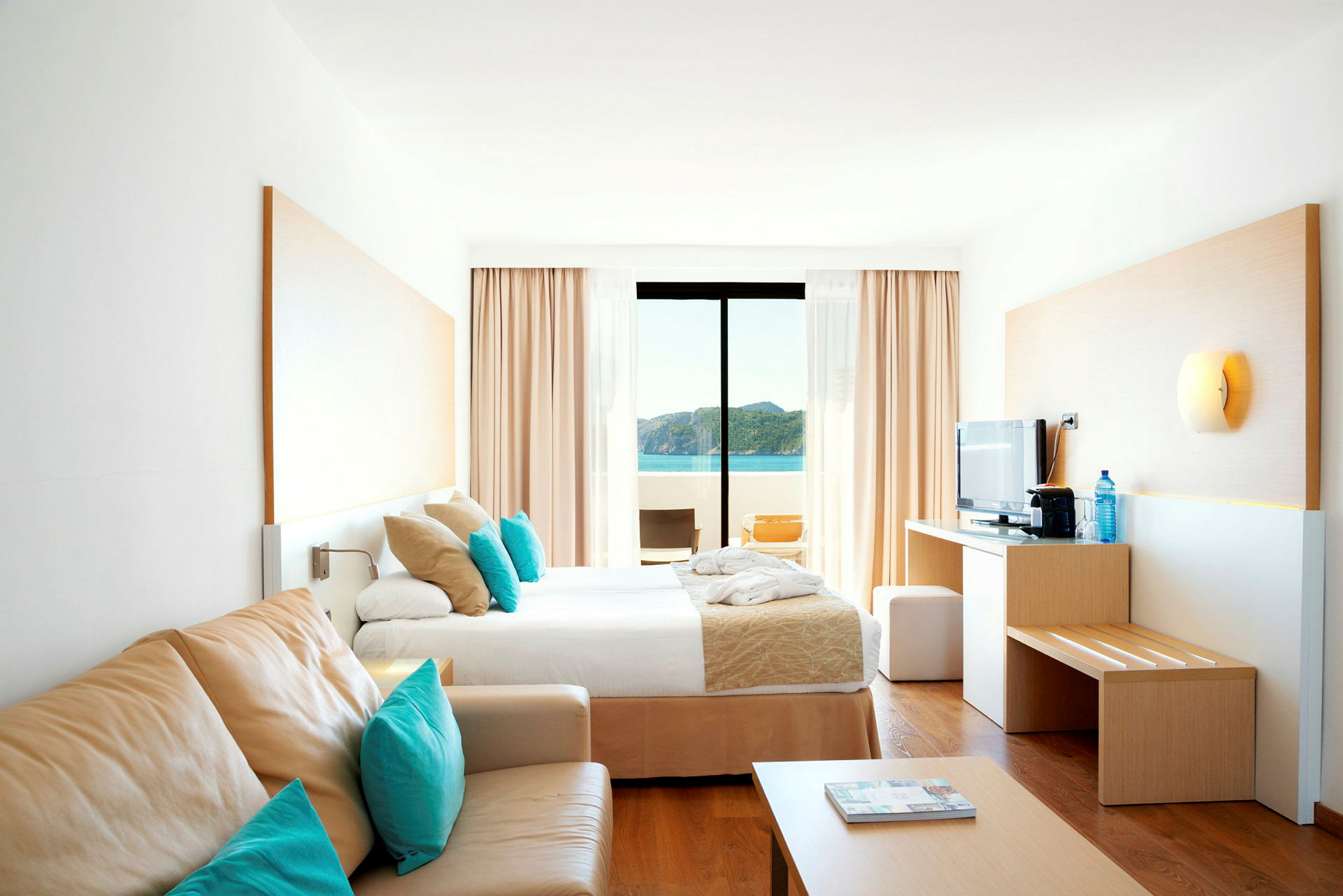 Et lyst hotellrom med en seng, sofa og utsikt over havet. (AI-generert tekst)