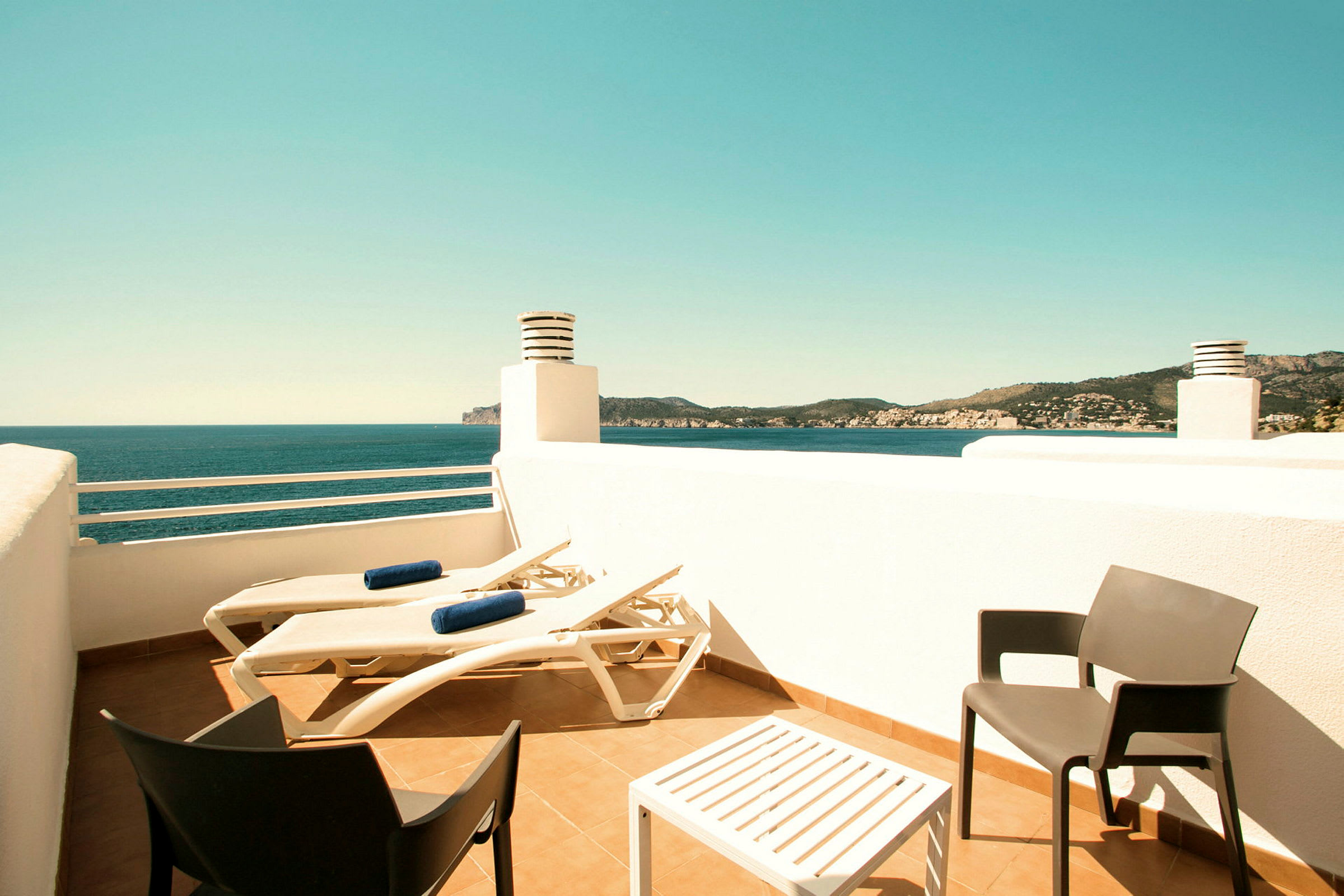 En terrasse med solsenger, et bord og to stoler samt utsikt over havet og omkringliggende landskap. (AI-generert tekst)