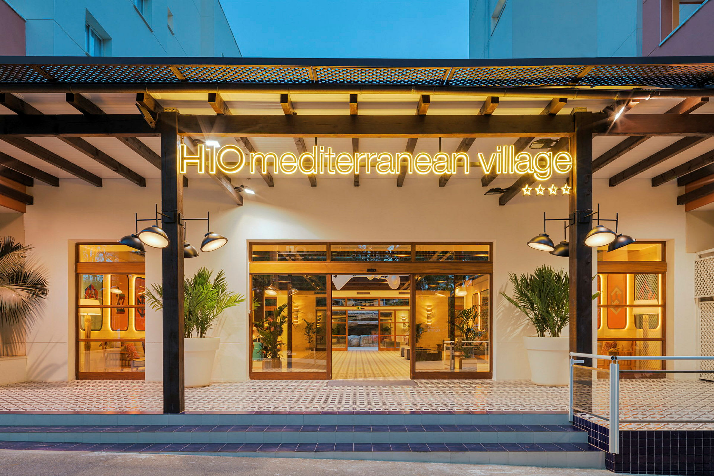 Inngangen til H10 Mediterranean Village, et hotell med opplyst skilt, store vinduer og moderne design. (AI-generert tekst)