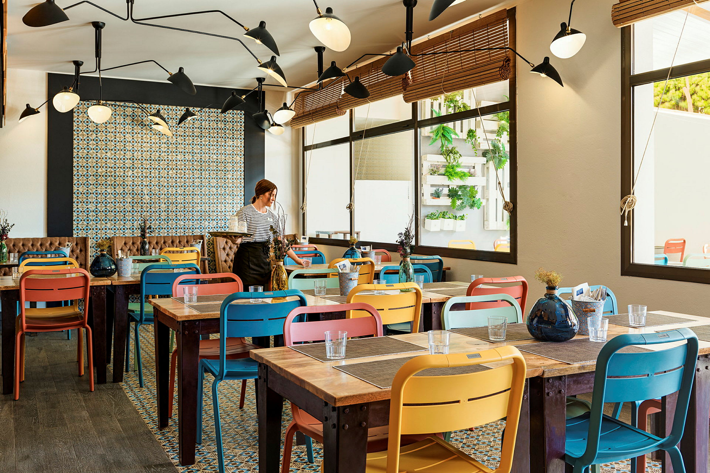 Innendørsbilde fra en restaurant med fargerike stoler og trebord. En person står ved et av bordene. (AI-generert tekst)
