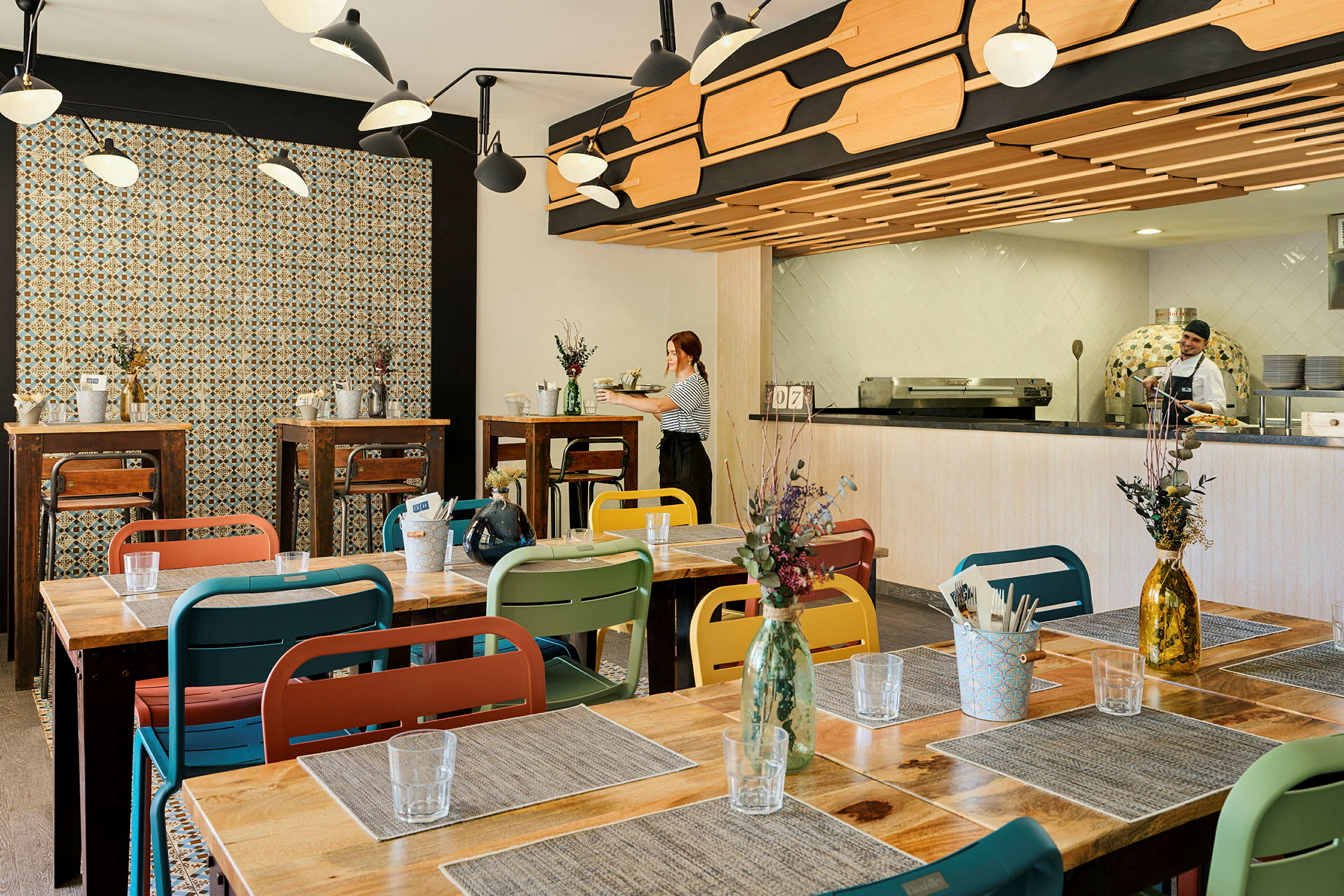 Bilde av et restaurantmiljø med fargerike stoler og trebord, dekorert med blomster i vaser. Veggene har mønstret flis og kjøkkenet er synlig i bakgrunnen. (AI-generert tekst)