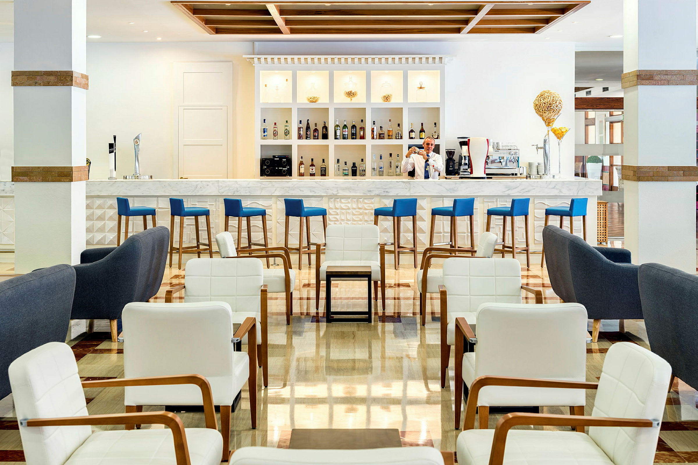 En moderne hotellbar med blå barkrakker, marmordisk og hvit innredning. Foran er en sittegruppe med hvite og grå lenestoler. (AI-generert tekst)