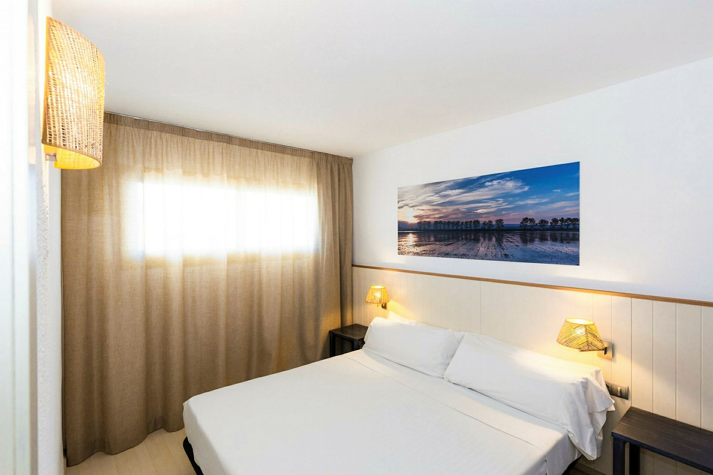 Et hotellrom med en seng dekket i hvitt sengetøy, en vegglampe og et kunstverk på veggen. Et vindu med elegante beige gardiner. (AI-generert tekst)