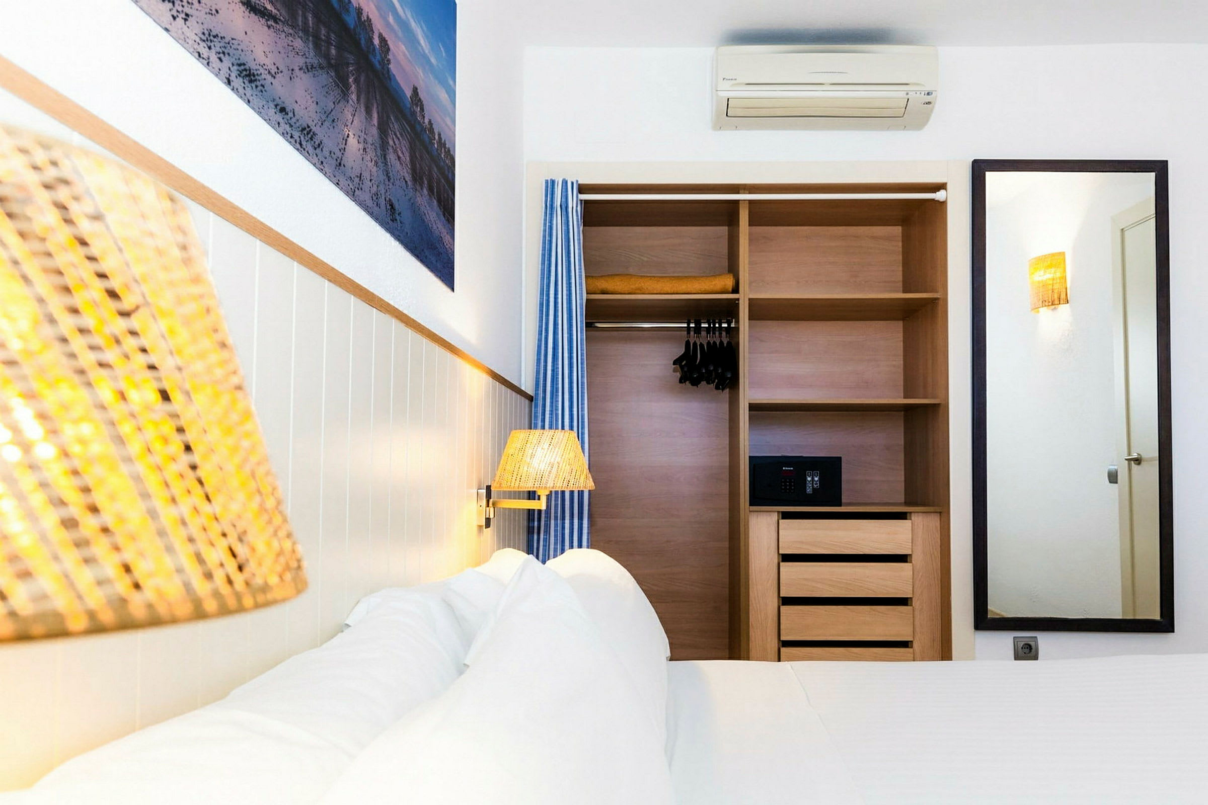 Et hotellrom med en seng og en innebygd garderobe. Garderoben er åpen og inneholder hyller, kleshengere og en safe. På veggen er det et speil og et maleri. (AI-generert tekst)