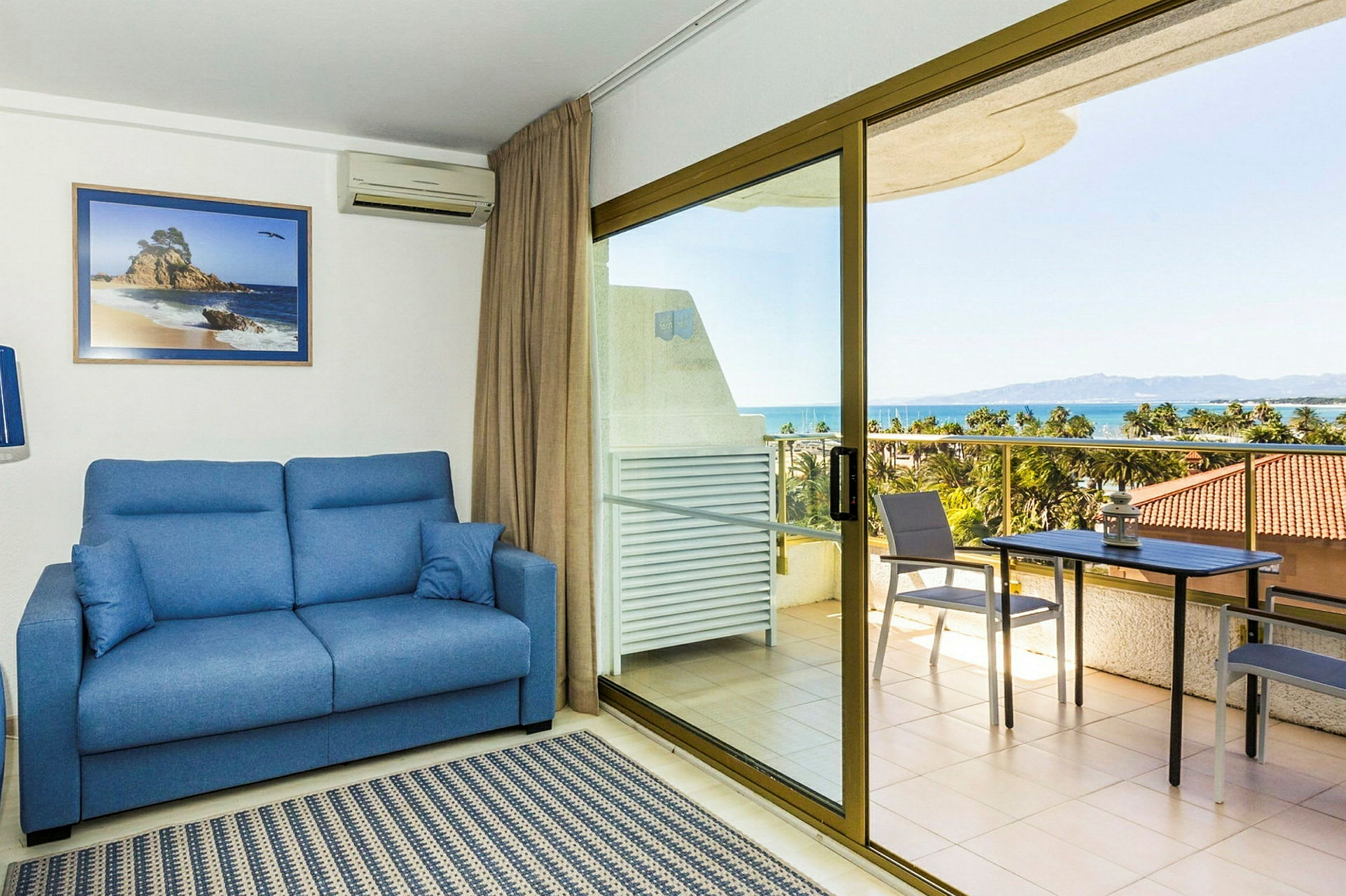 Et rom med en blå sofa, teppe og et glasstakvindu ut mot en terrasse. Terrassen har bord, stoler og utsikt over havet. (AI-generert tekst)