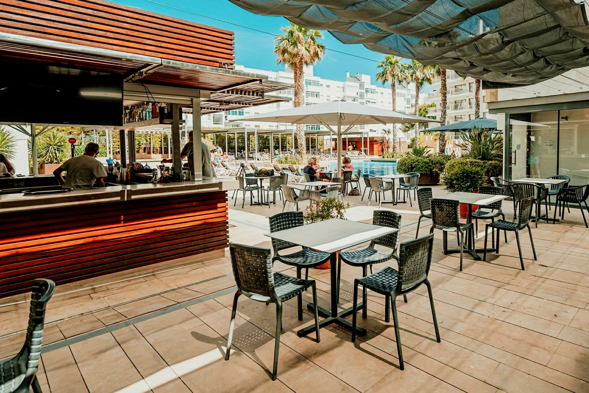 En bassengnær restaurant med sitteplasser utendørs og en serveringsdisk, omgitt av bygninger og palmer. (AI-generert tekst)