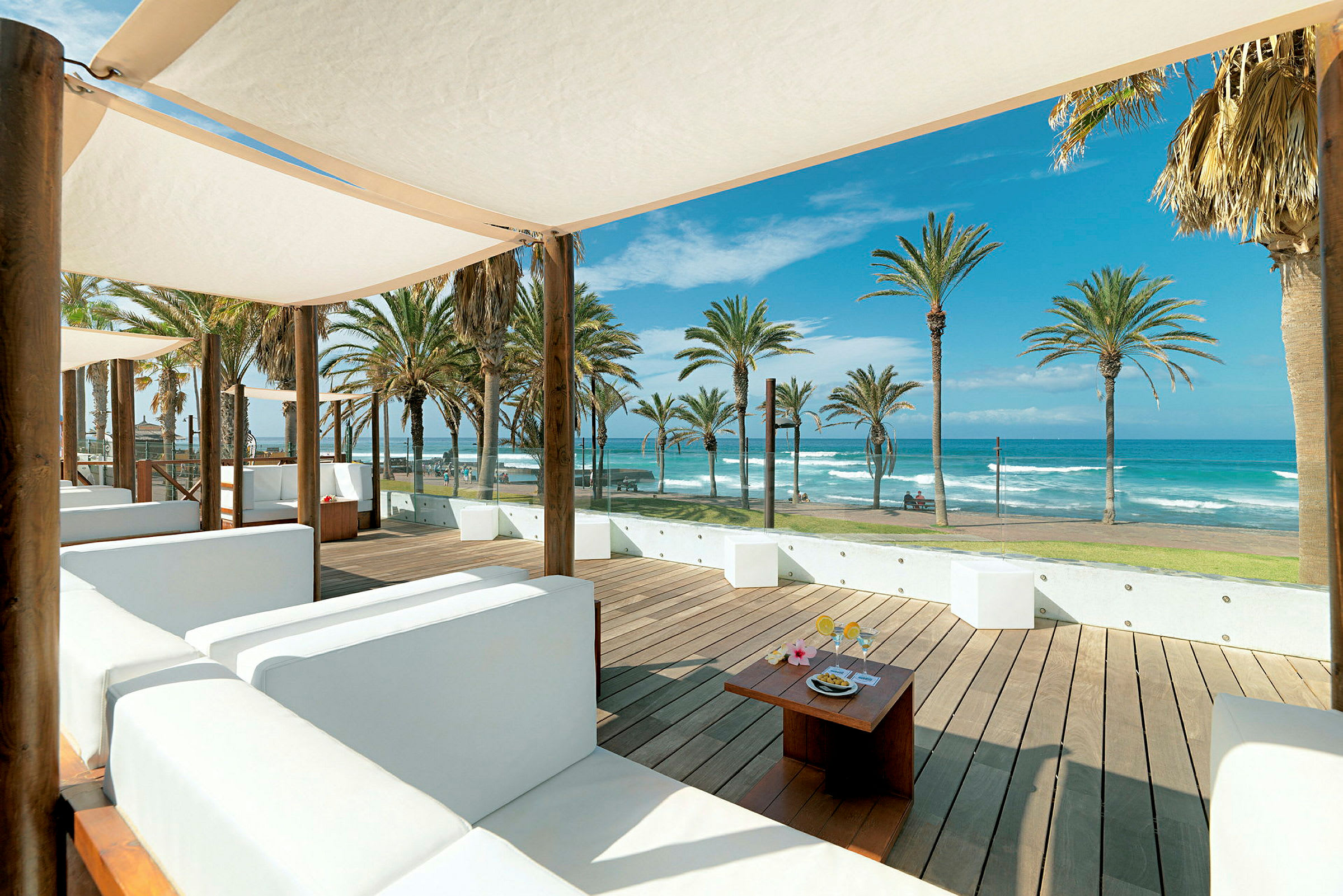En utendørslounge med hvite sofaer og parasoller med utsikt over stranden og havet. (AI-generert tekst)