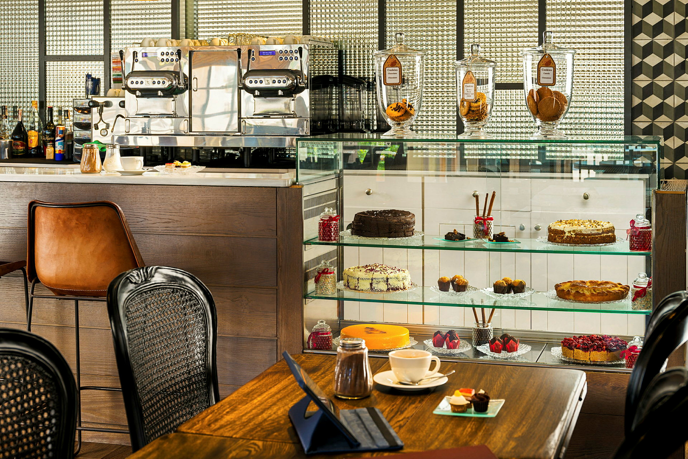 Et bilde av en kafé med kaker og bakverk i en glassmonter, en espressomaskin og elegante kaffekopper på en bordflate. (AI-generert tekst)