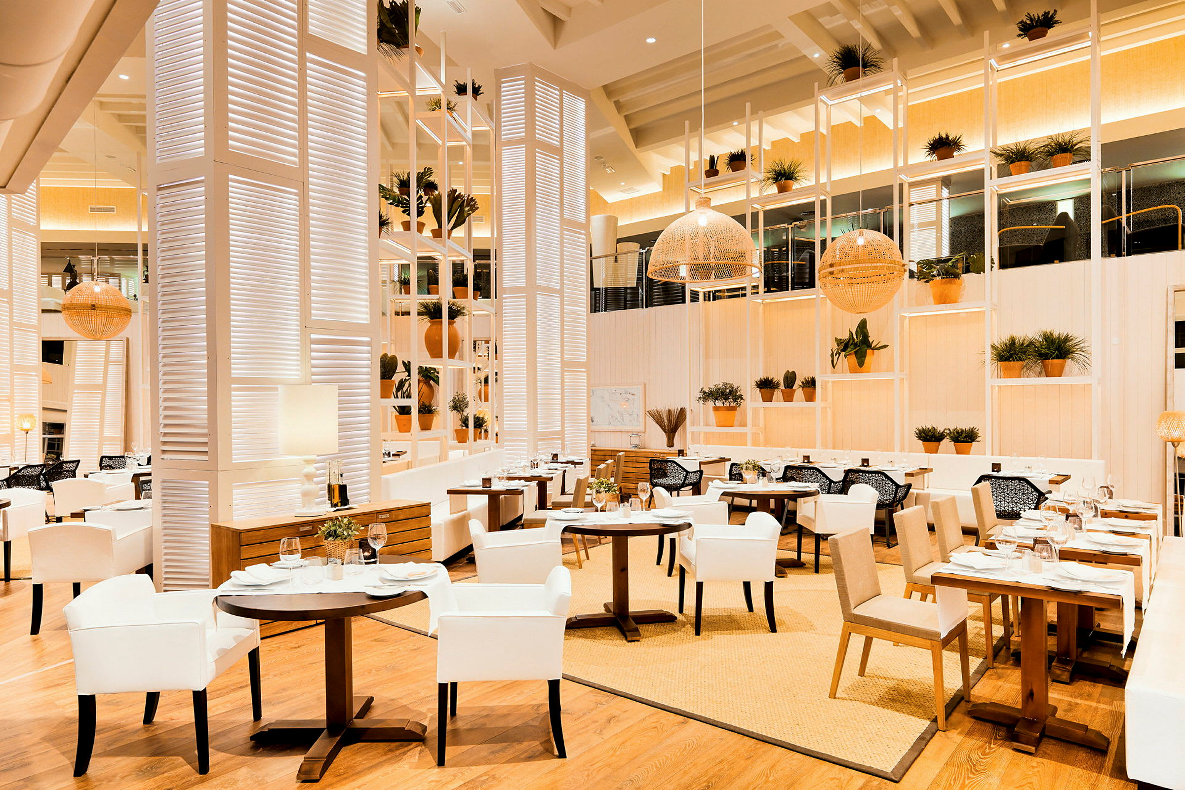 En elegant restaurant med lys innredning, hvite og beige møbler samt trematerialer. Potteplanter pryder hyllene og bordene. (AI-generert tekst)
