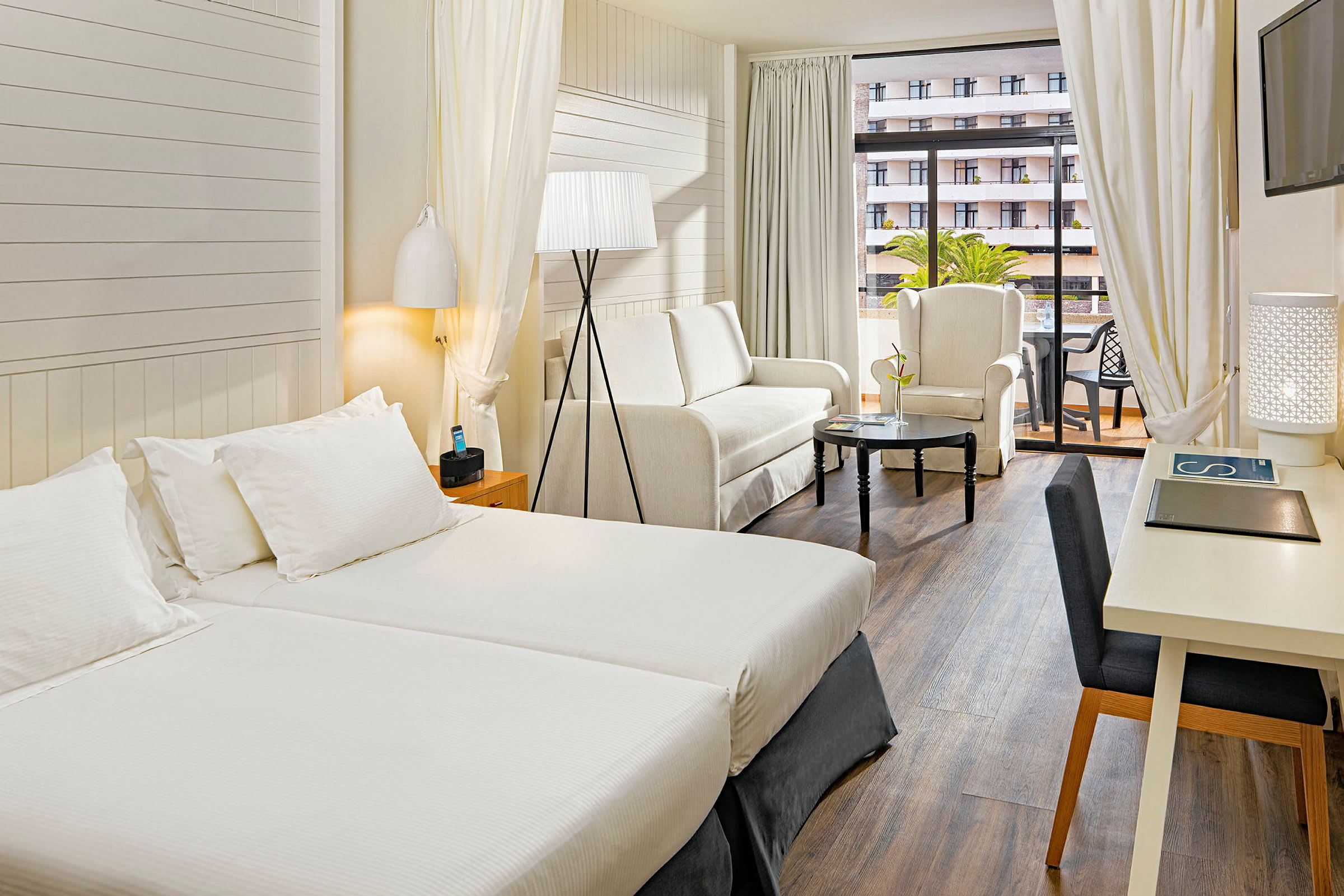 Et hotellrom med to enkeltsenger, en sittegruppe med sofa og lenestol samt et bord. Det er også et skrivebord og en balkong. (AI-generert tekst)