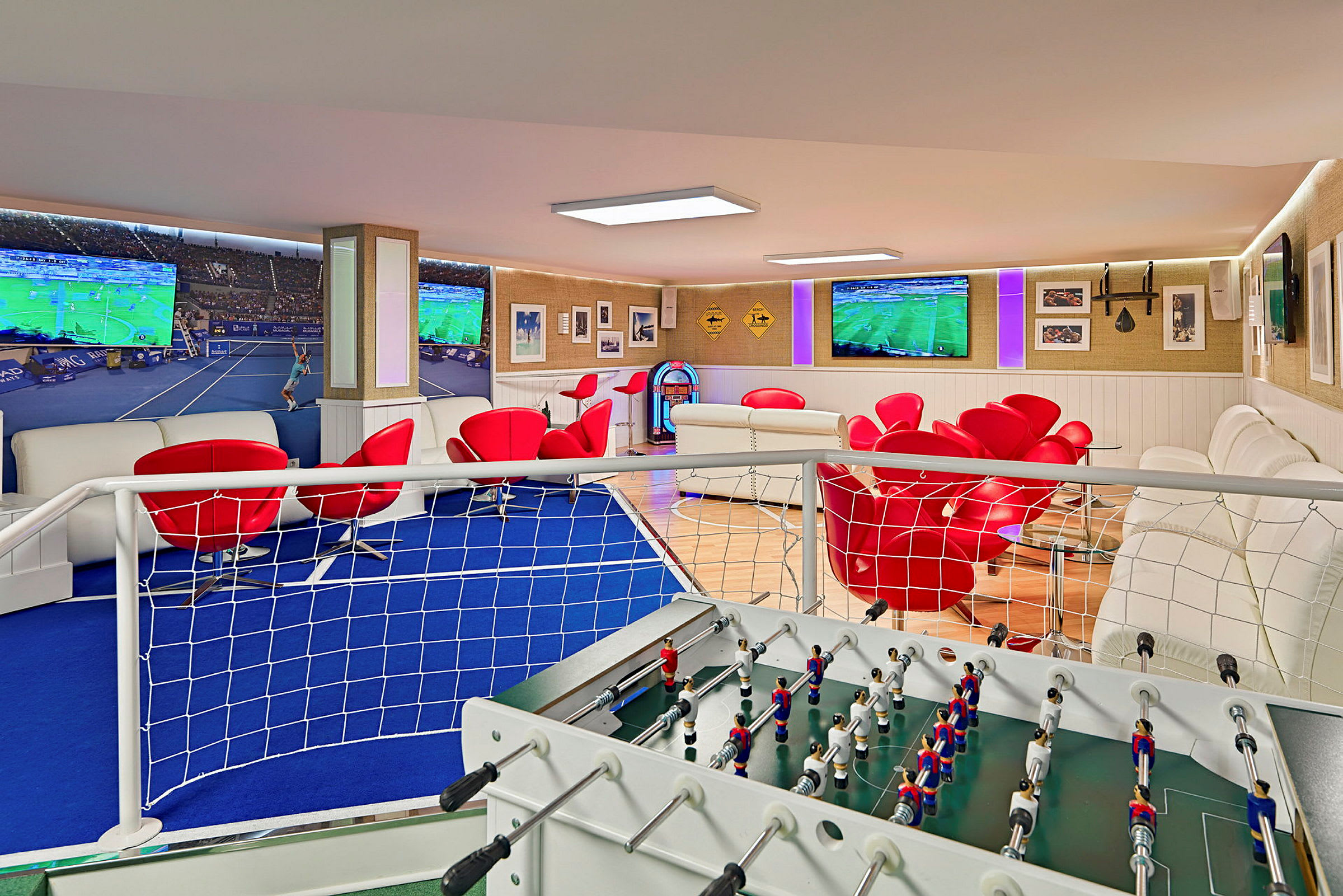 Et rom med et foosballbord i forgrunnen, røde stoler, hvite sofaer og flere store TV-skjermer som viser sport. (AI-generert tekst)