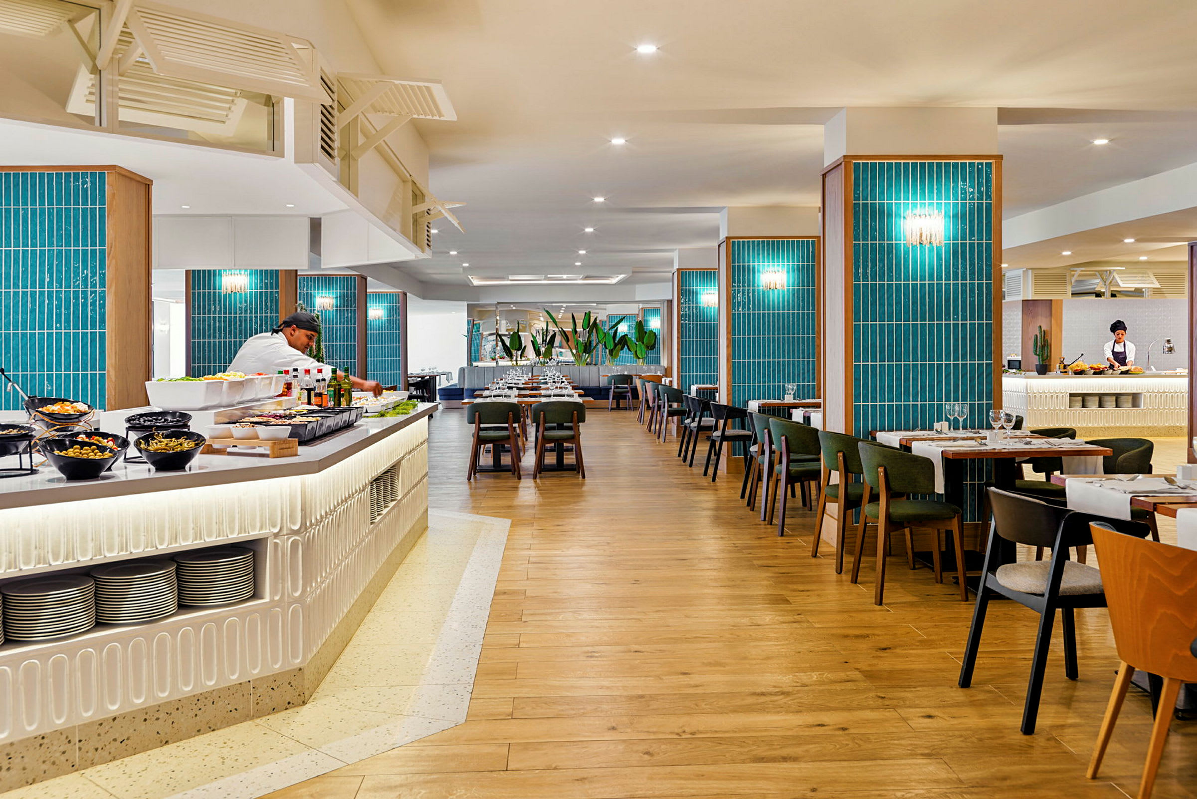 En romslig restaurant med tregulv, blå veggdekorasjoner og bord med stoler. En buffédisk med salater og drikkevarer er synlig i forgrunnen, og en kokk jobber bak disken. (AI-generert tekst)