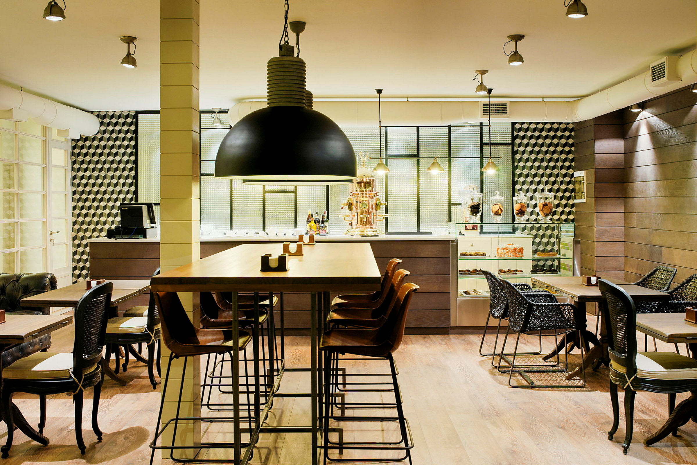 En moderne kafé med høye og lave bord, samt stoler i forskjellige design. Interiøret er stilrent med detaljer som svart hengelampe, geometriske veggmønster og en disk med bakverk. (AI-generert tekst)