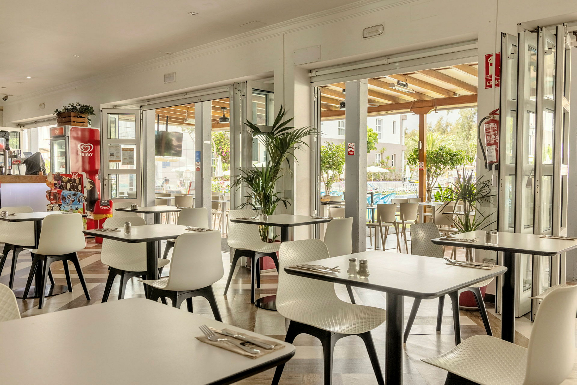 En restaurant med hvite stoler og bord, åpen mot en terrasse og omgivelser med grøntområder. (AI-generert tekst)