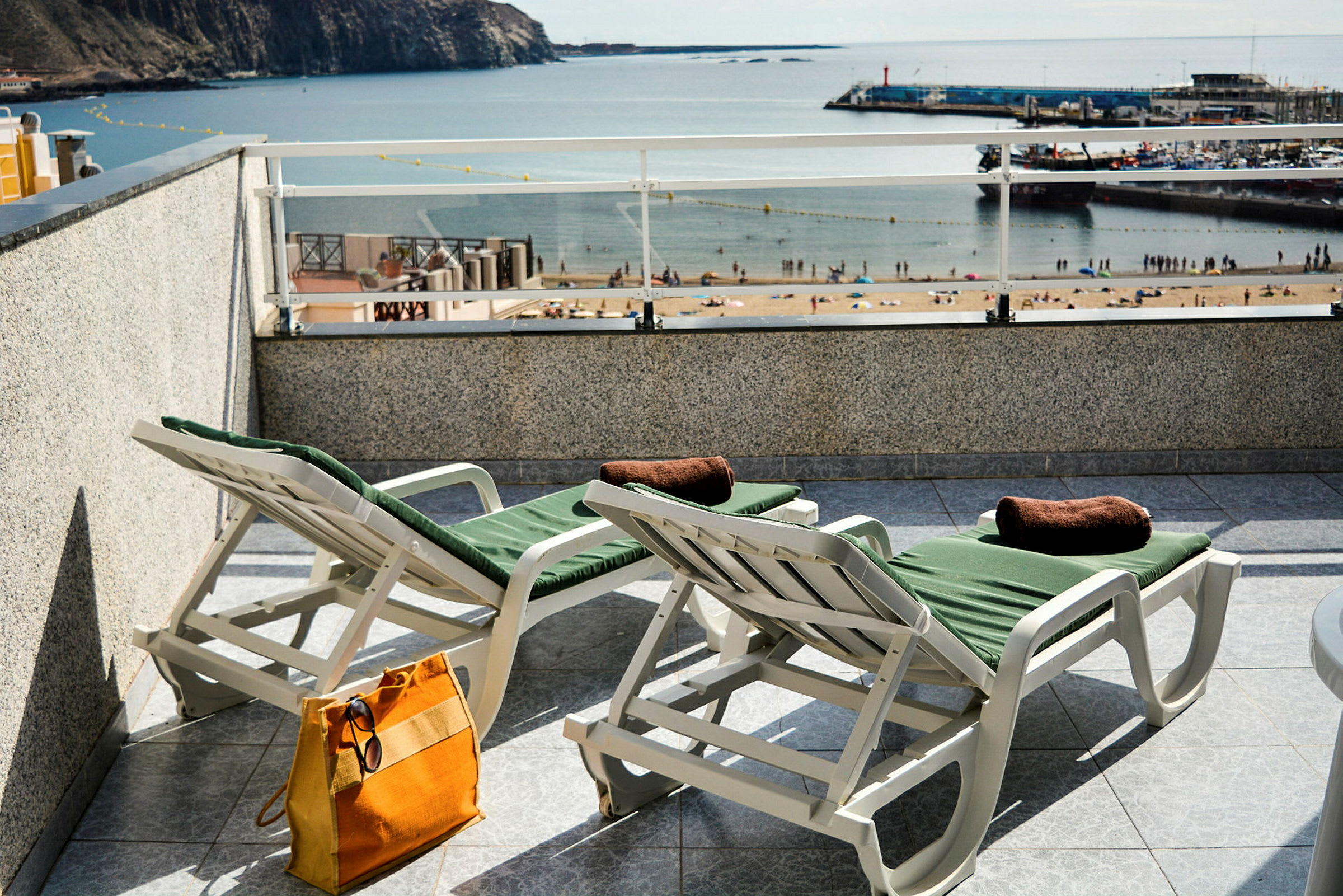 To solsenger med grønne puter på en terrasse med utsikt over en strand og havn. (AI-generert tekst)