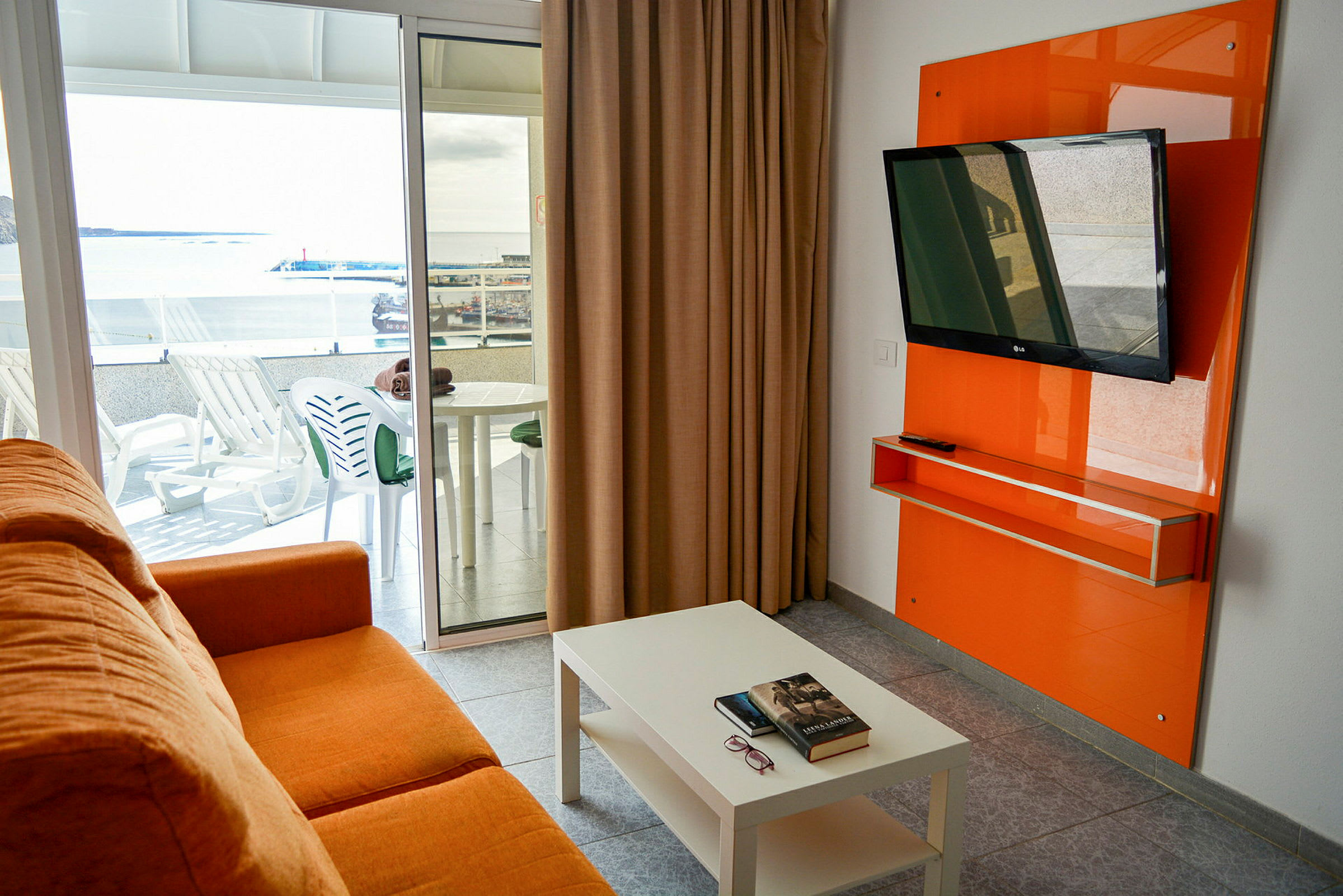 En oransje sofa i et rom med et hvitt bord, en flat-TV på veggen og en balkong med utemøbler samt utsikt over et havneområde. (AI-generert tekst)