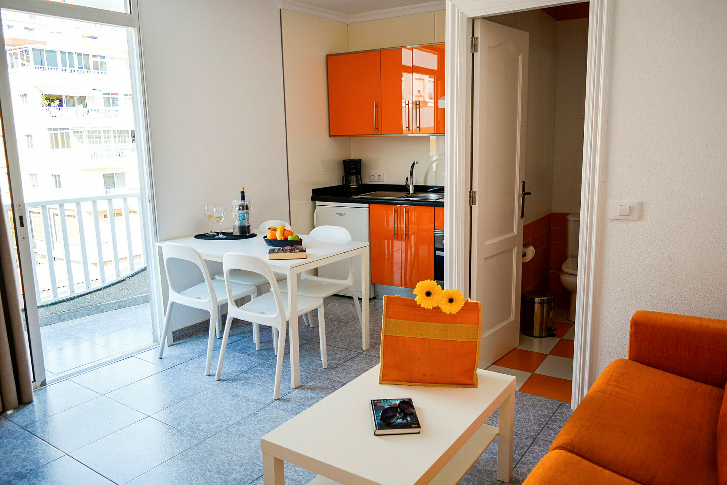 En liten leilighet med oransje kjøkkeninnredning, en hvit spiseplass og en sofa. (AI-generert tekst)