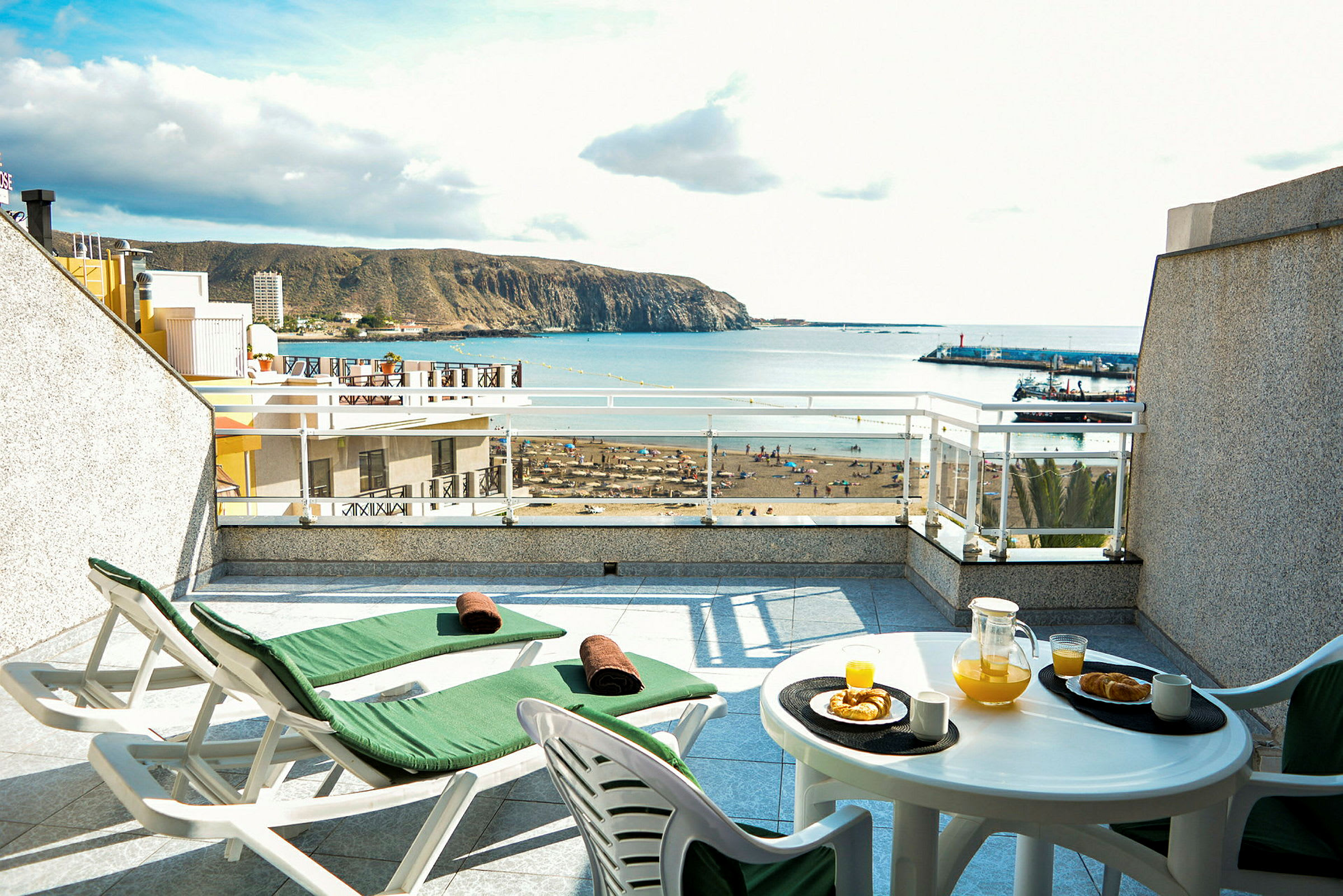Terrasse med to solsenger og et bord med appelsinjuice og bakverk, havutsikt i bakgrunnen. (AI-generert tekst)