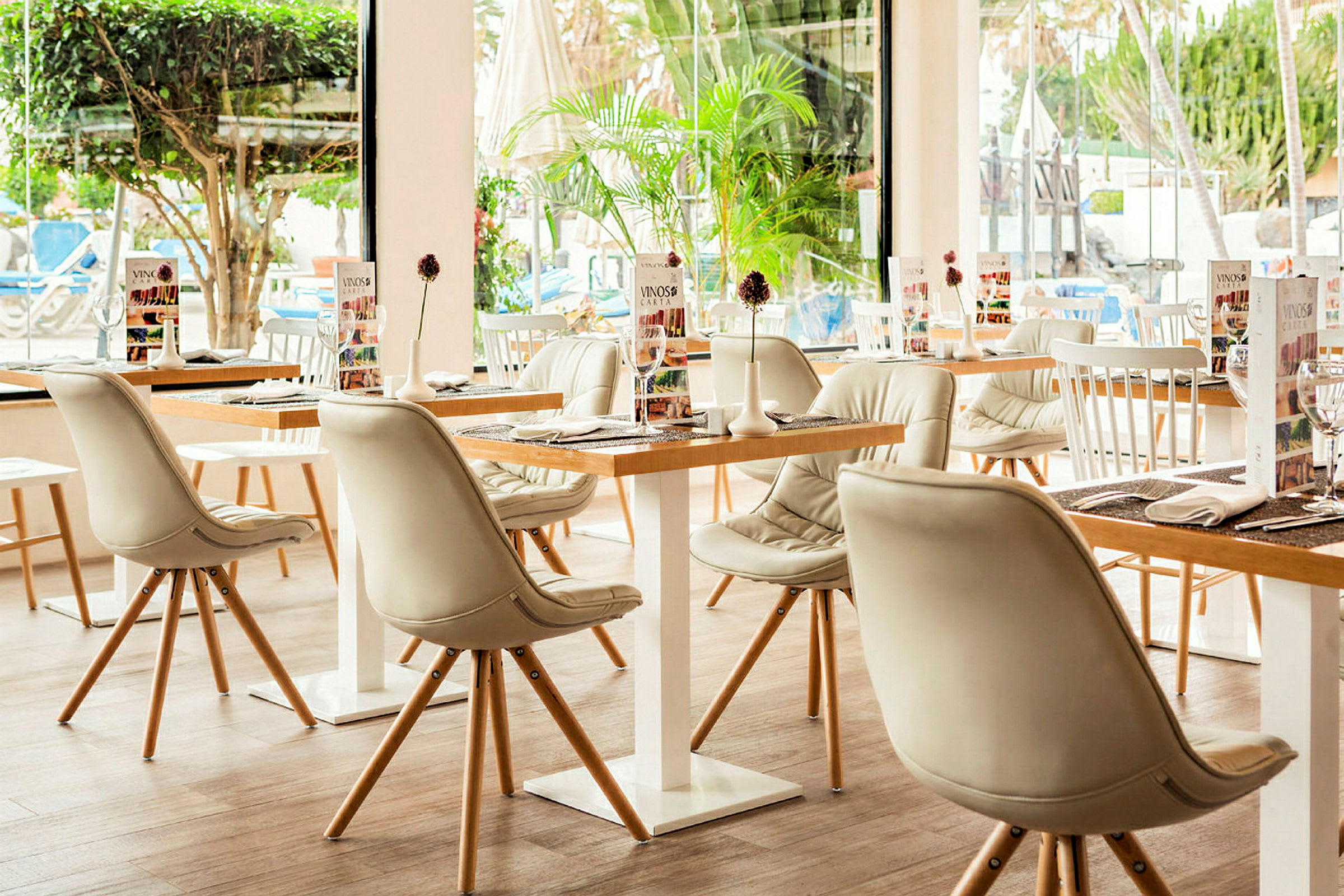 Restaurant med hvite stoler og tremøbler, med glassvinduer og planter i bakgrunnen. (AI-generert tekst)