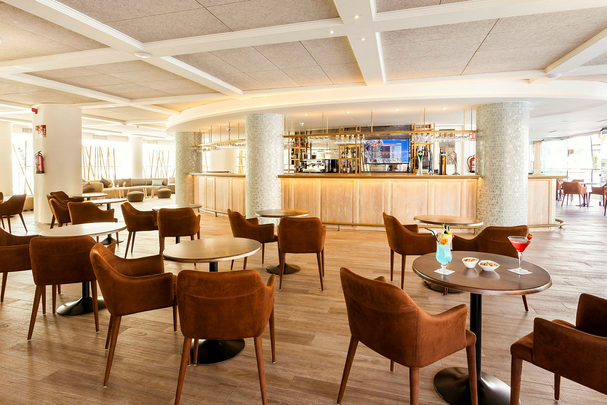 En hotellbar med elegante brune stoler og glassbord, tre drinker og småretter servert, samt en lys og moderne atmosfære. (AI-generert tekst)