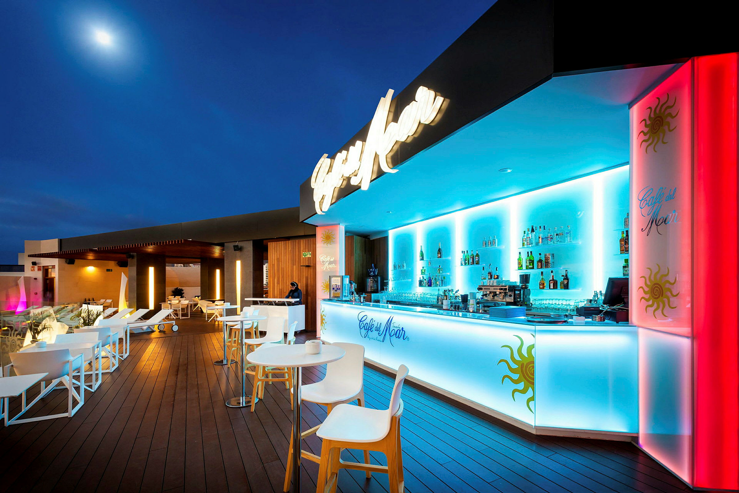 Bilde av en moderne bar med blå belysning, hvite møbler og en hevet terrasse med mørkt tregulv. (AI-generert tekst)