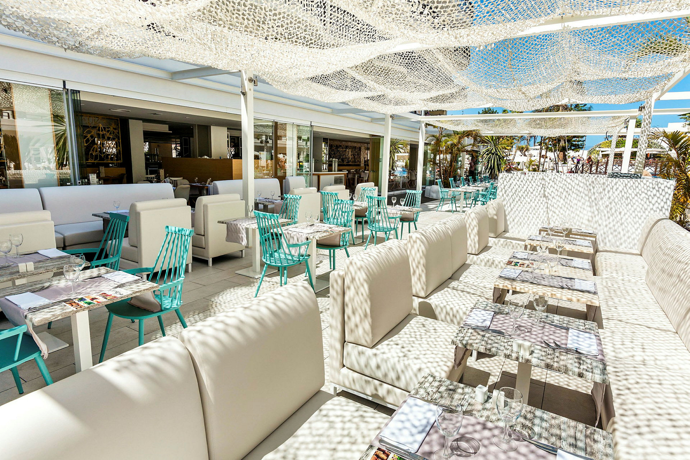 En utendørs restaurant med hvite sittemøbler og turkise stoler, dekorert med lysgjennomslippende nett som skaper skyggemønstre. (AI-generert tekst)
