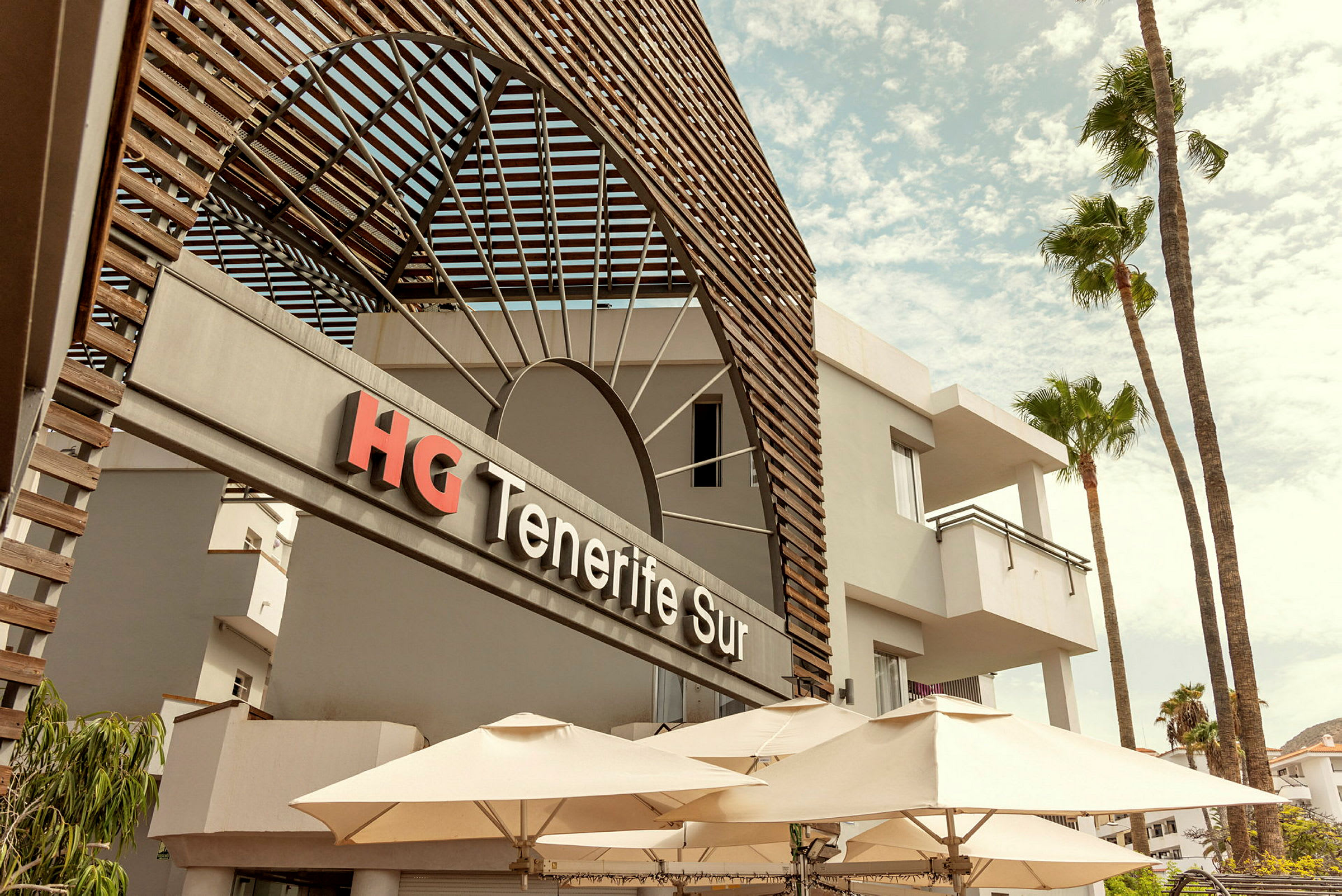 Forsiden av HG Tenerife Sur-hotellet med parasoller og palmer. (AI-generert tekst)