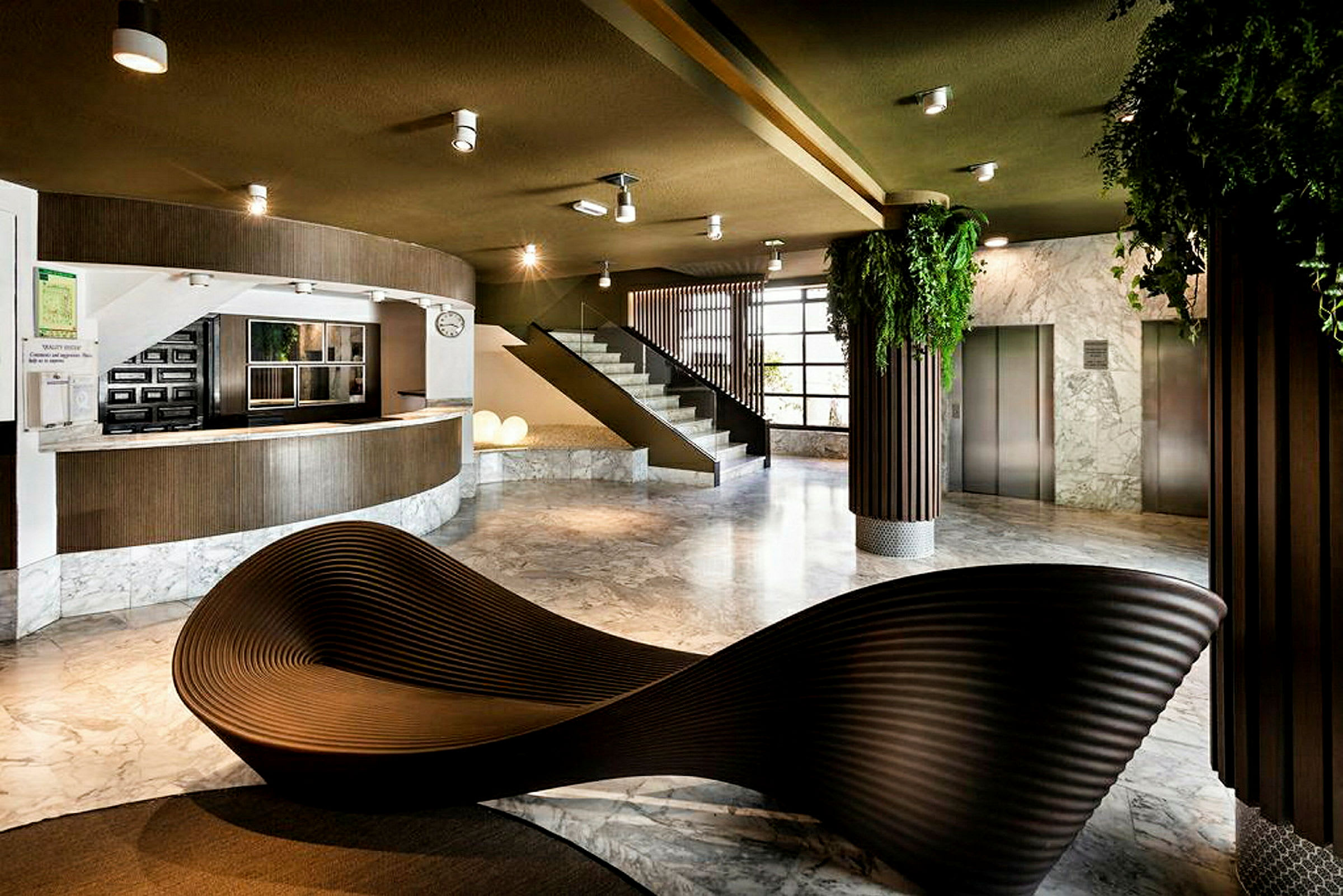 Et moderne lobbyområde med resepsjonsdisk, trapp og heiser. Interiøret inkluderer en skulpturell sittemøbel og grønne planter. (AI-generert tekst)