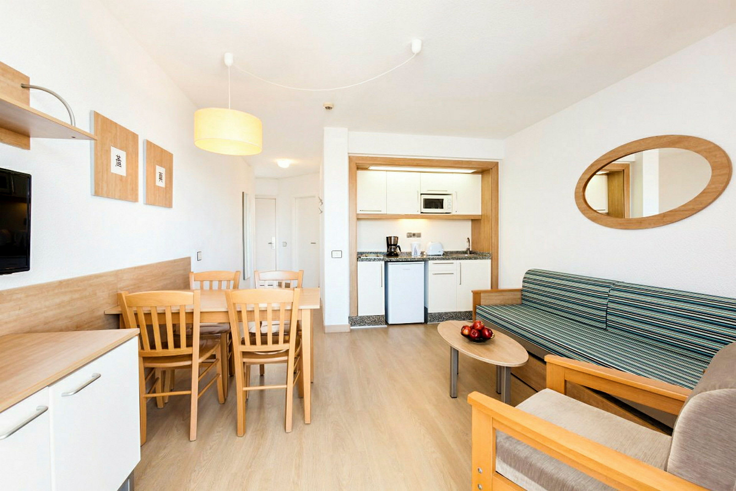 Et moderne hotellrom med en liten kjøkkenkrok, en spiseplass med bord og stoler samt en sittegruppe med sofa og salongbord. (AI-generert tekst)