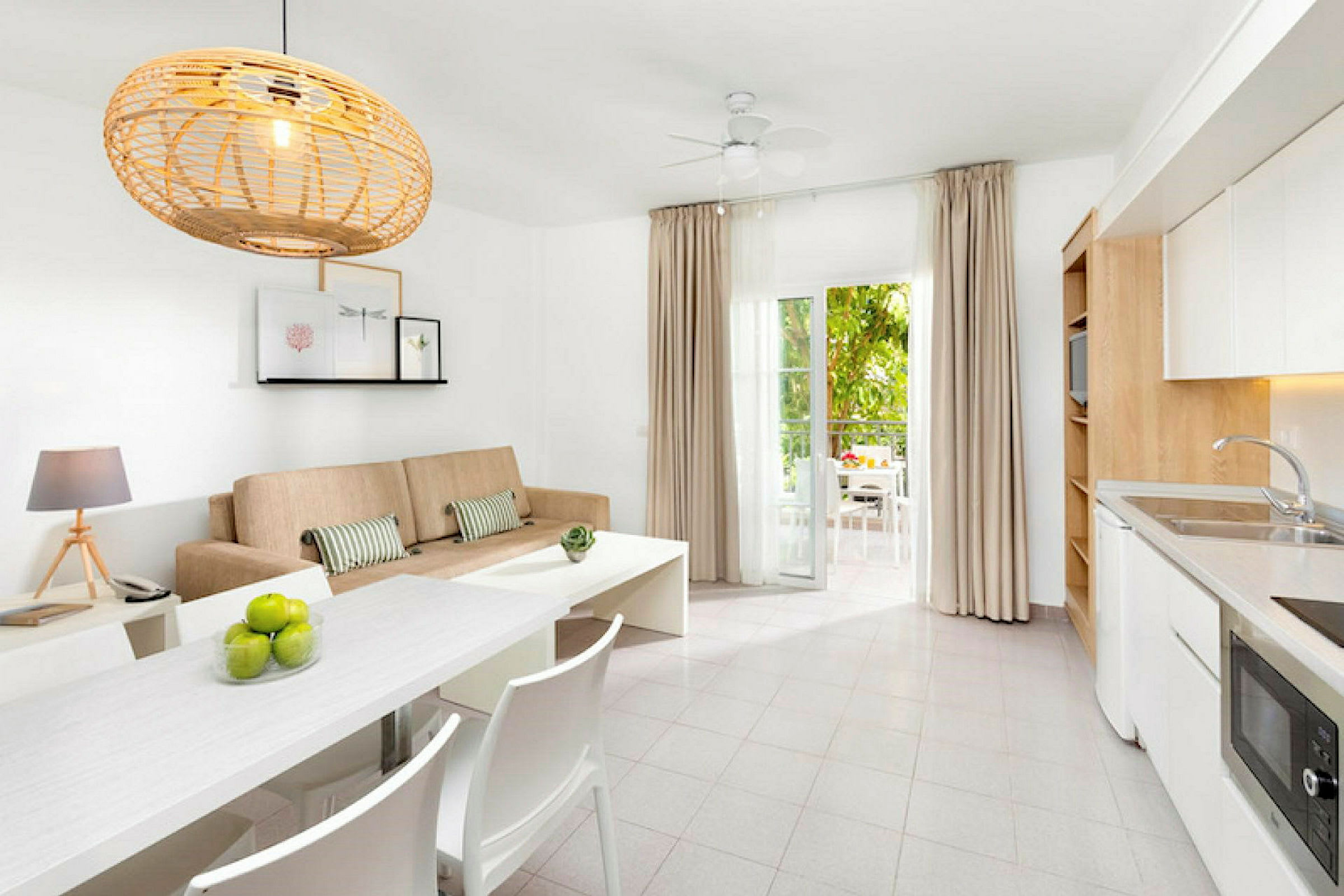 Et lys og moderne innredet rom med en beige sofa, hvitt spisebord og kjøkken, samt åpne dører til en møblert balkong. (AI-generert tekst)