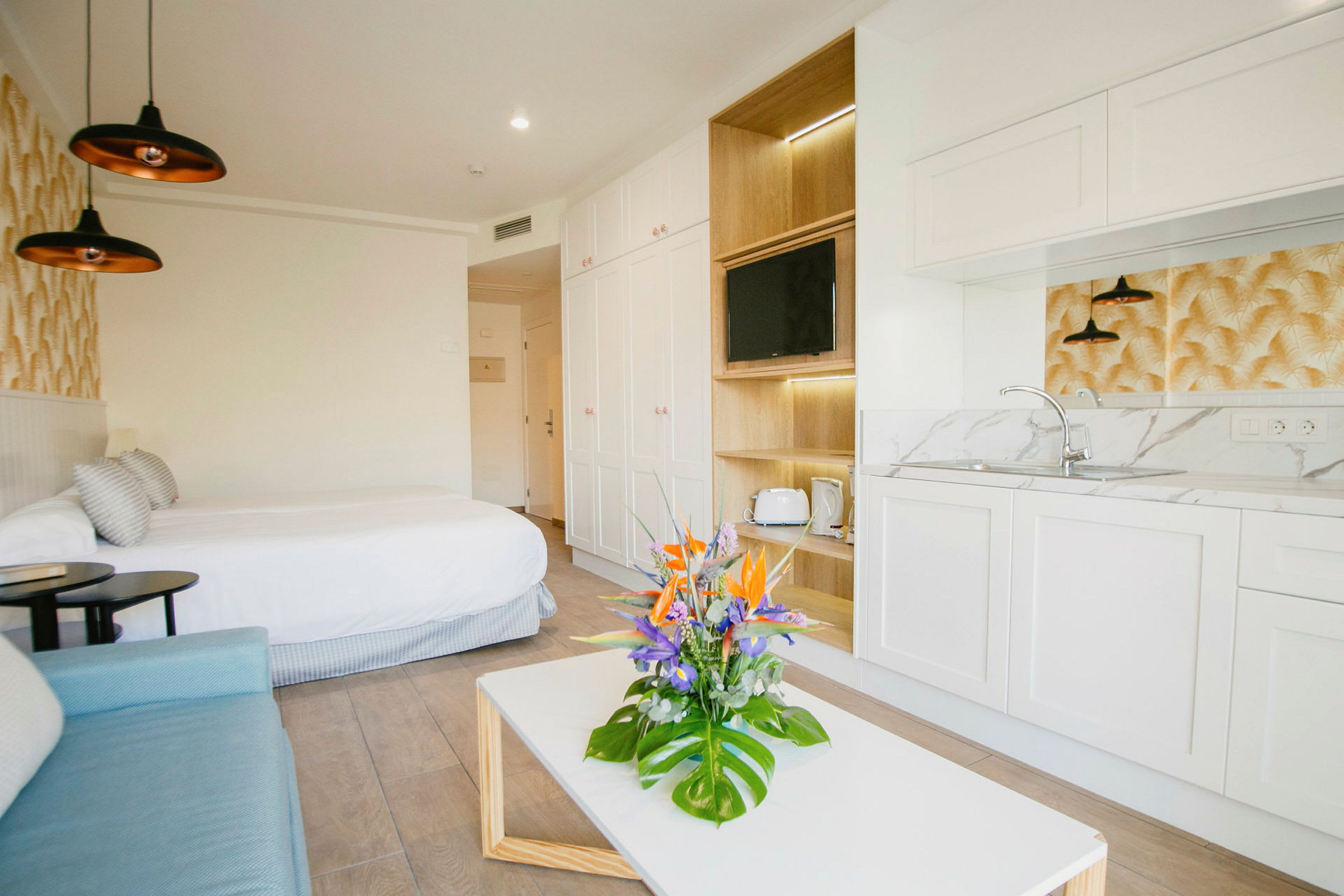 Et moderne hotellrom med en dobbeltseng, sofa, lite kjøkken og et bord med en blomsterbukett. (AI-generert tekst)